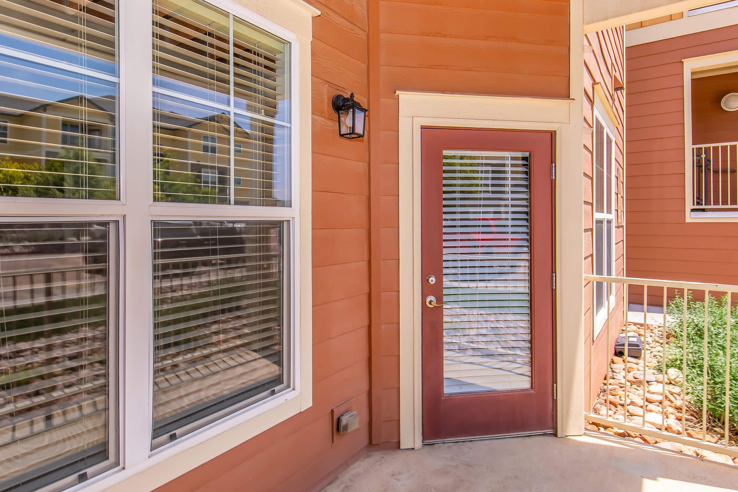 A close-up view of a brown exterior wall featuring a door with a glass insert and blinds. The door is framed by two large windows with white frames. The setting includes a small porch area with a railing and pebbles along the ground, surrounded by greenery. Bright sunlight casts natural shadows.