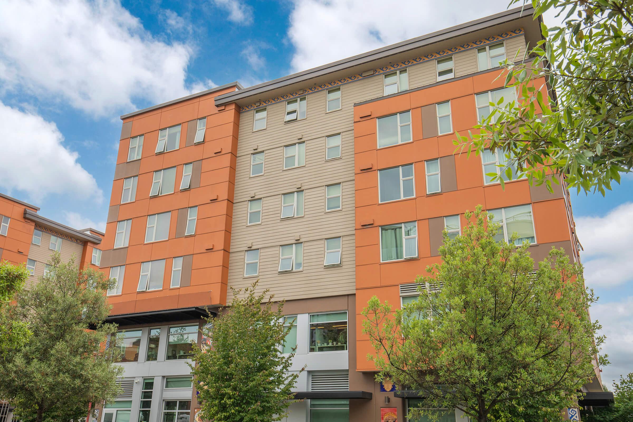 A modern multi-story residential building featuring a mix of orange and beige siding, with large windows and greenery around the base. The sky is partly cloudy, providing a bright atmosphere.