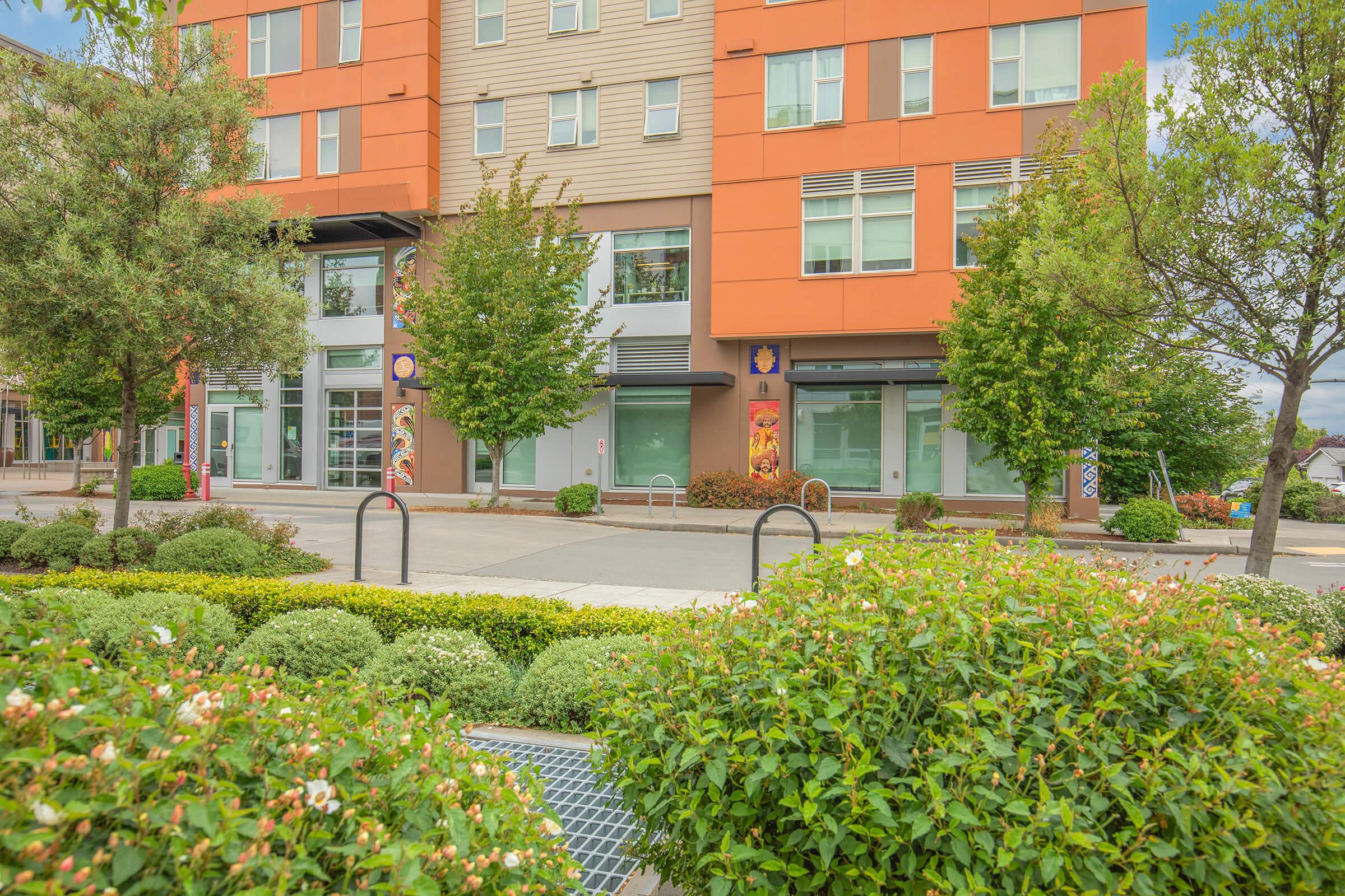 A modern building with orange and gray exterior, featuring large windows and greenery in front. Flowering bushes and small trees line the walkway, while decorative banners hang on the walls. The scene is set in an urban environment, showcasing a clean and inviting space.