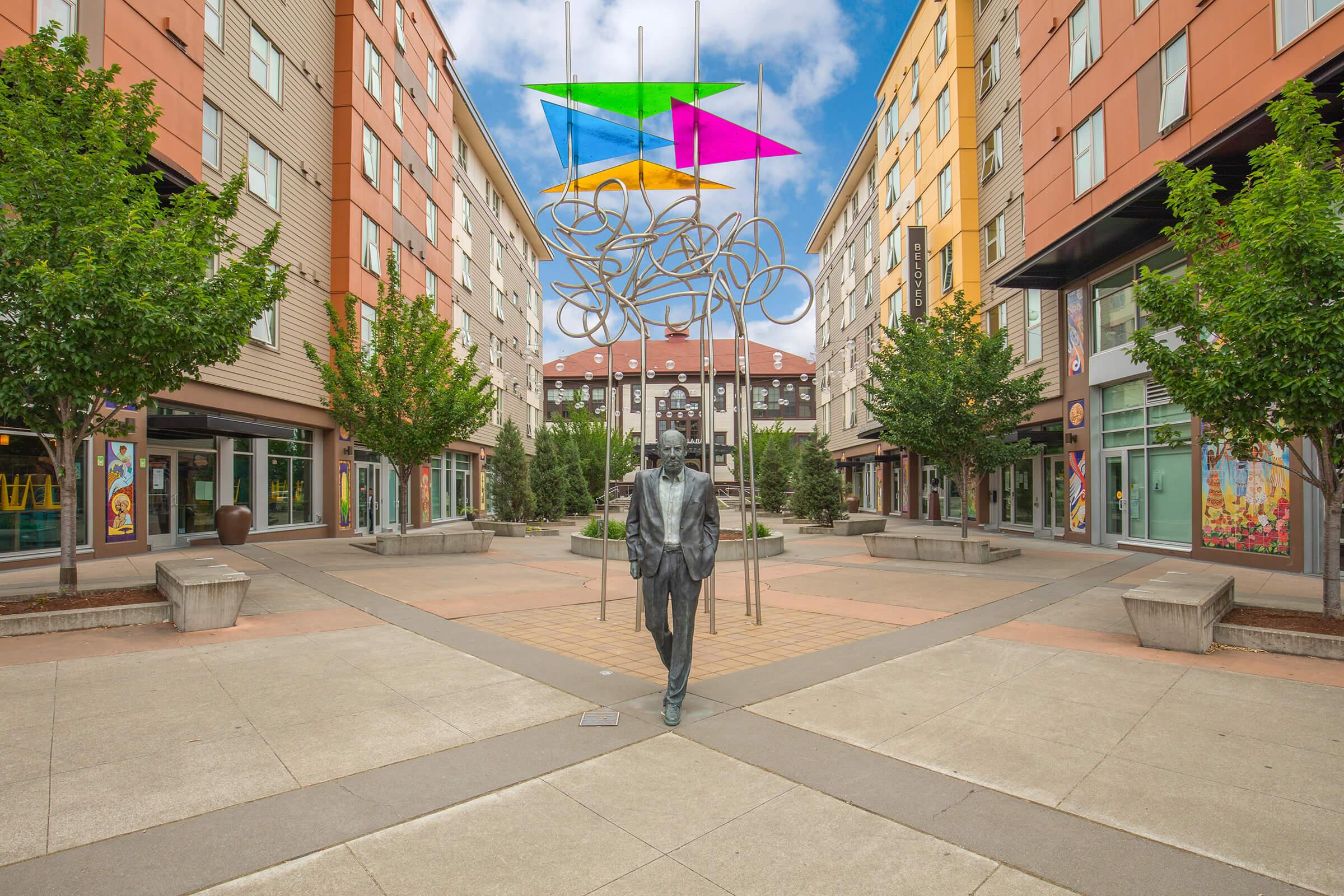 A modern urban plaza featuring a striking metal sculpture of a man walking, surrounded by colorful suspended shapes in shades of green, pink, and yellow. The area is lined with trees and contemporary buildings, with shops visible in the background under a blue sky.