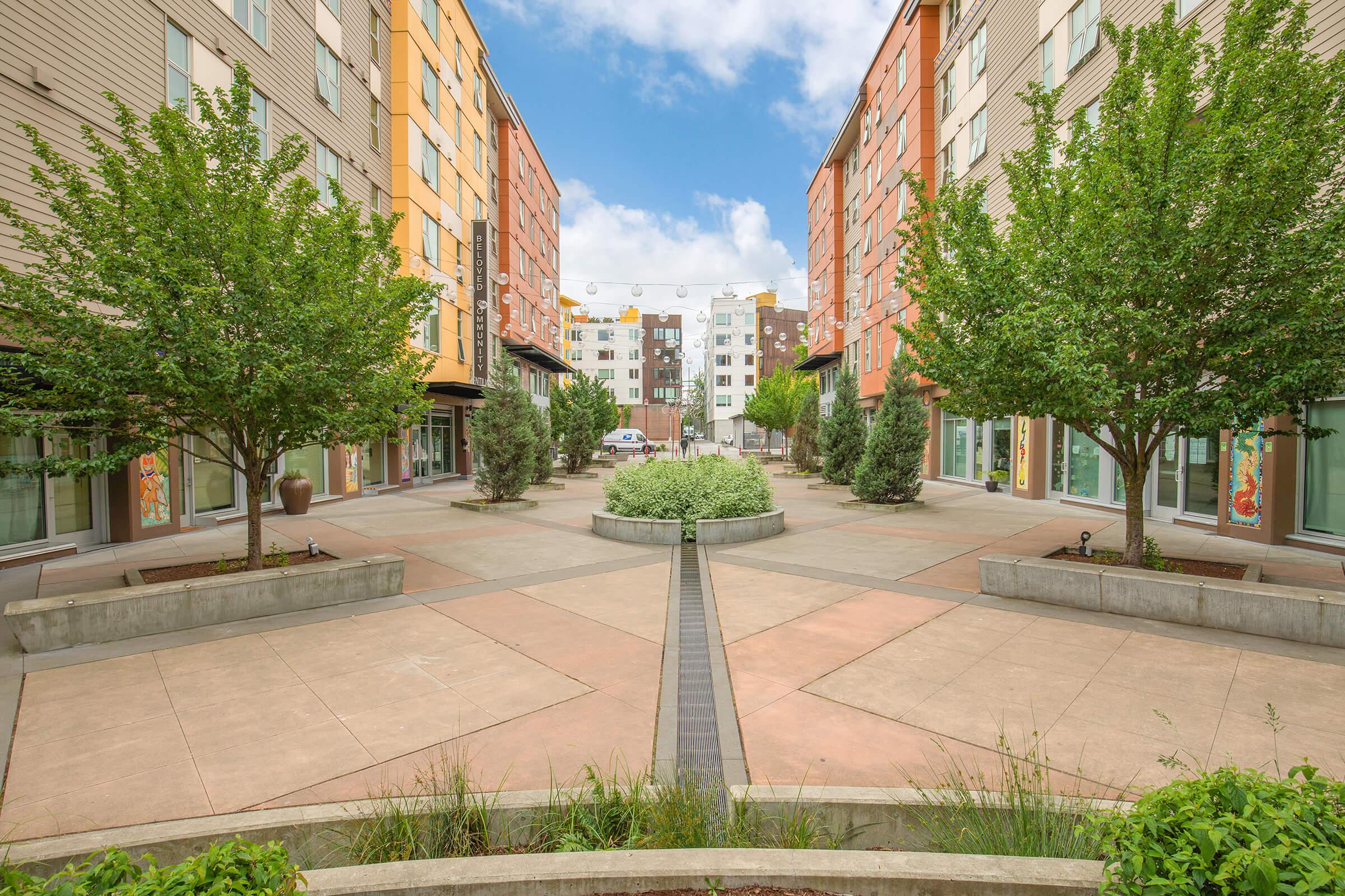 A landscaped courtyard surrounded by modern apartment buildings, featuring neatly arranged trees and plants. A concrete pathway runs through the center, leading to a grassy area. The sky is partly cloudy, with colorful building exteriors visible on either side.
