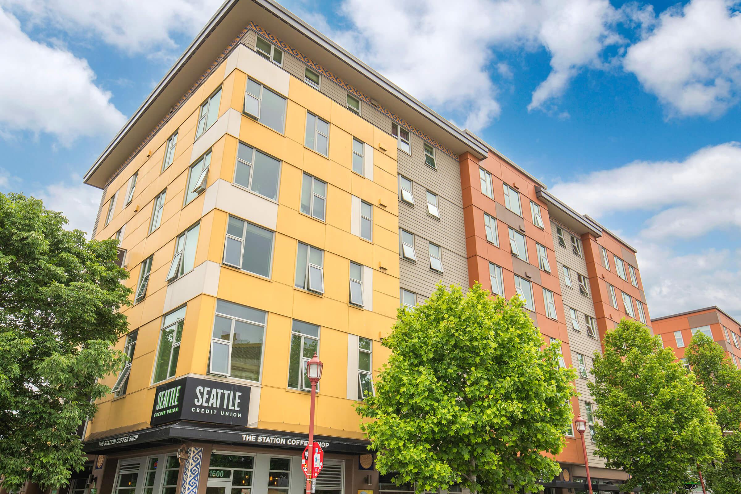 A modern multi-story building with a mix of yellow and orange exterior, featuring retail spaces on the ground floor, including a Seattle Credit Union sign and a coffee shop. There are trees and clear blue skies in the background, giving a vibrant urban feel.