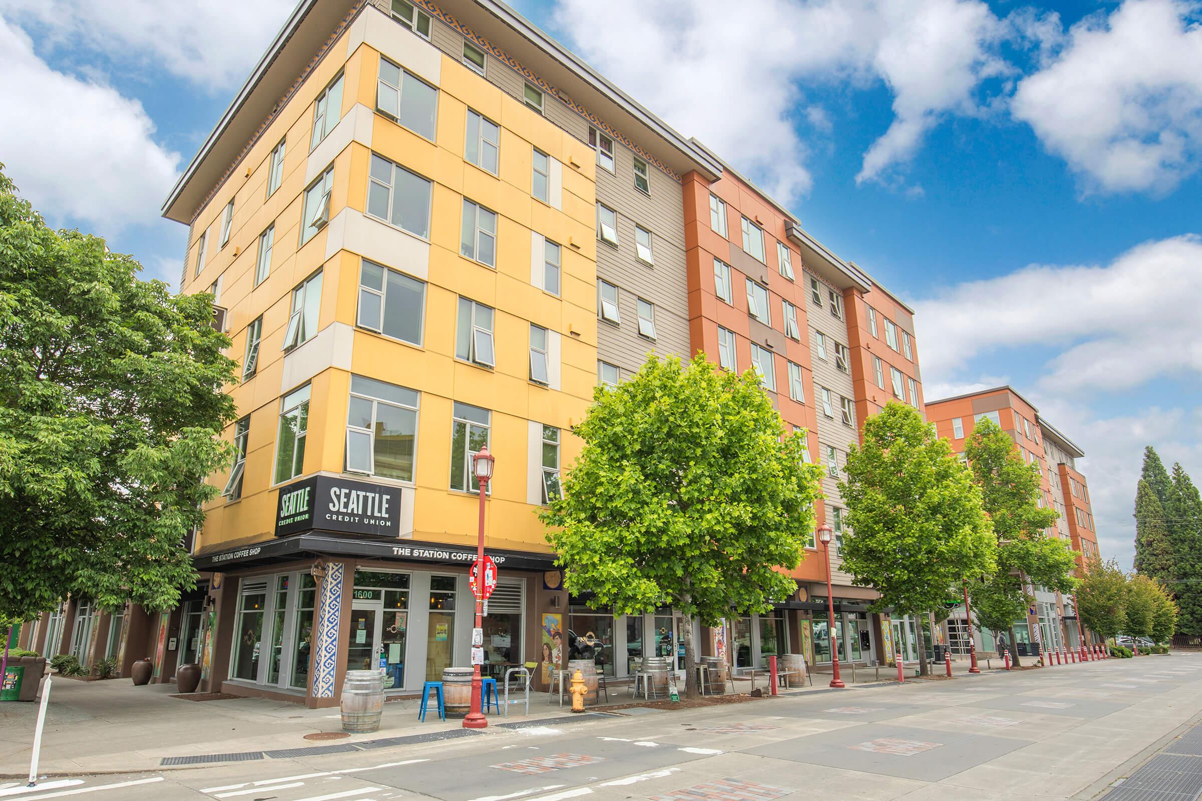 A multi-story building with a modern design featuring a mix of yellow and brown facades. The ground floor hosts a cafe named "Seattle Coffee." Surrounding the building are green trees and outdoor seating areas, with a blue sky scattered with clouds above.