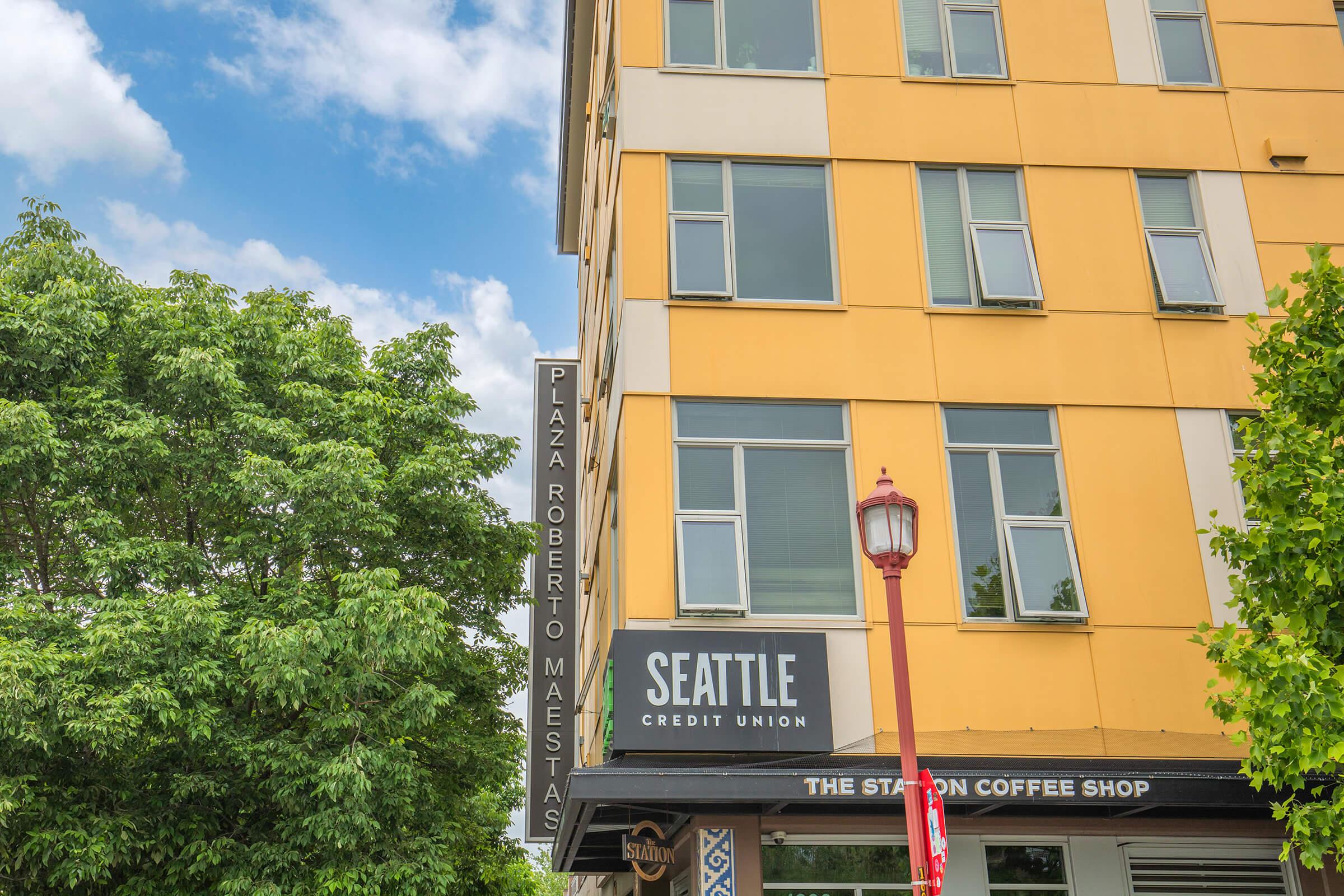 A corner of a building featuring a sign for "Seattle Credit Union" and "Plaza Roberto Maestas." Below, there is a visible entrance to "The Station Coffee Shop." Lush green trees surround the area under a partly cloudy sky.