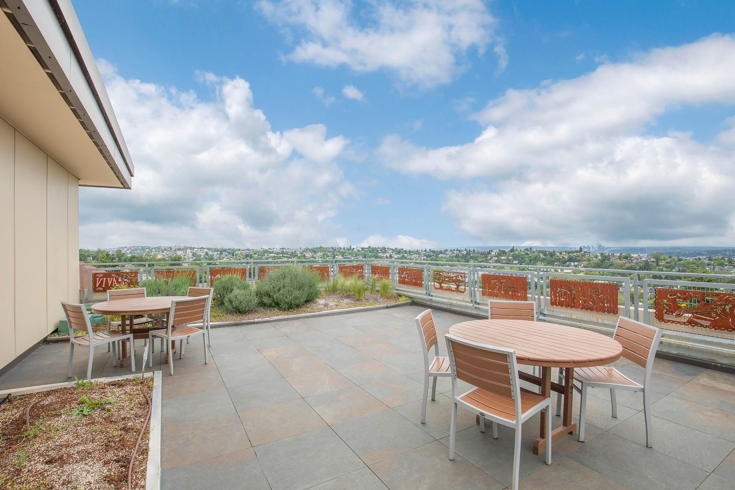 A rooftop terrace with circular wooden tables and metal chairs, surrounded by greenery. A decorative metal railing features intricate designs, offering a panoramic view of cityscape and cloudy blue sky in the background. The setting feels open and inviting, perfect for outdoor gatherings.