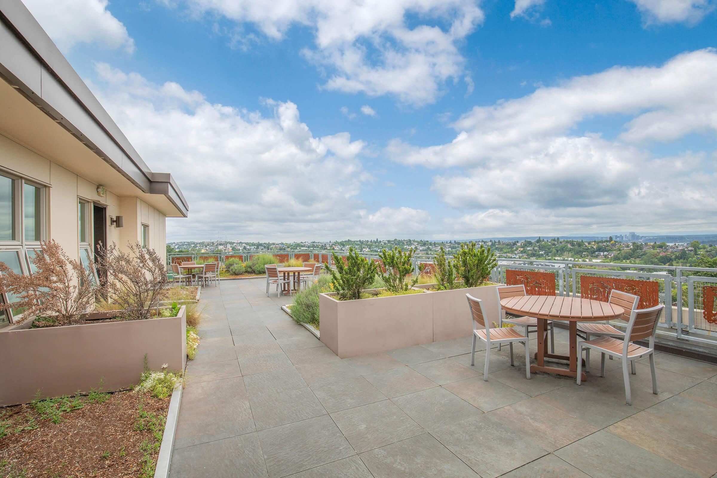 Rooftop terrace featuring a view of a city skyline under a partly cloudy sky. The area is equipped with wooden tables and chairs, surrounded by green plants in modern planters. The flooring is made of large stone tiles, creating a spacious and inviting outdoor setting.