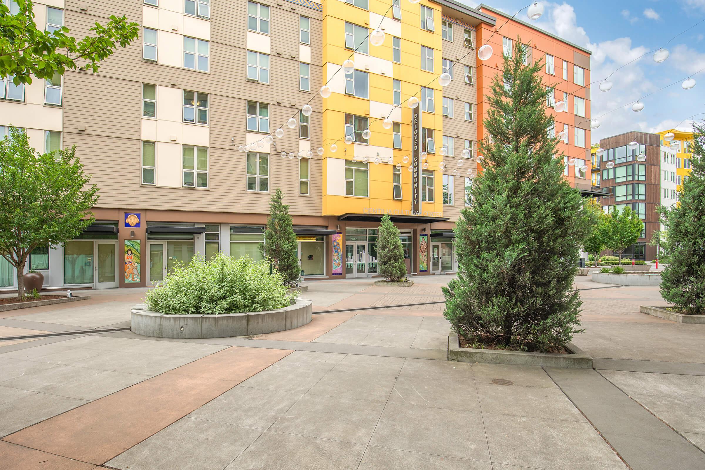 A modern outdoor plaza featuring greenery, stylish buildings with colorful facades, and a paved area. String lights are visible above, and there are planters with shrubs and small trees, creating a welcoming urban space. The scene is bright and inviting, ideal for community gatherings.