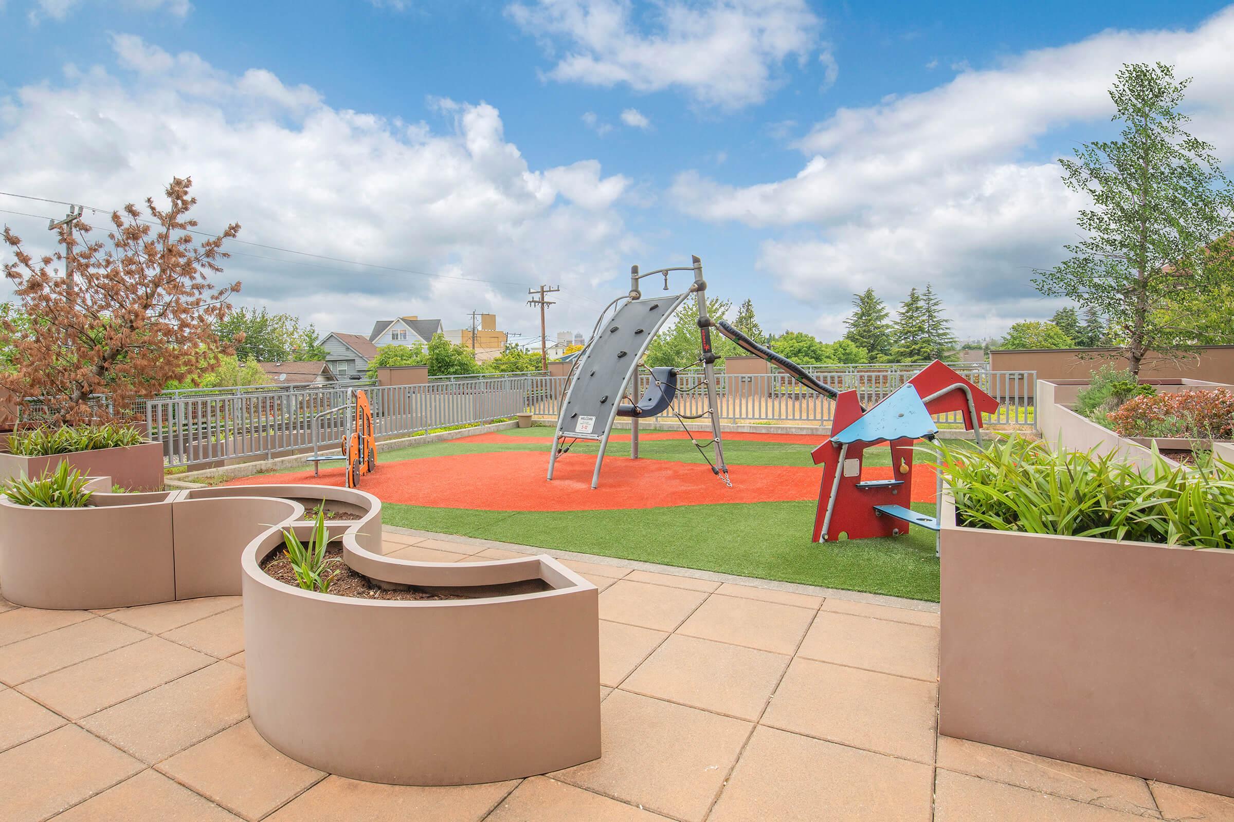 Playground area featuring a swing set, slide, and small playhouse, surrounded by green grass and decorative plants. Bright blue sky with fluffy clouds overhead. The flooring is a tan color, and there are fence barriers around the space, offering a safe environment for children.