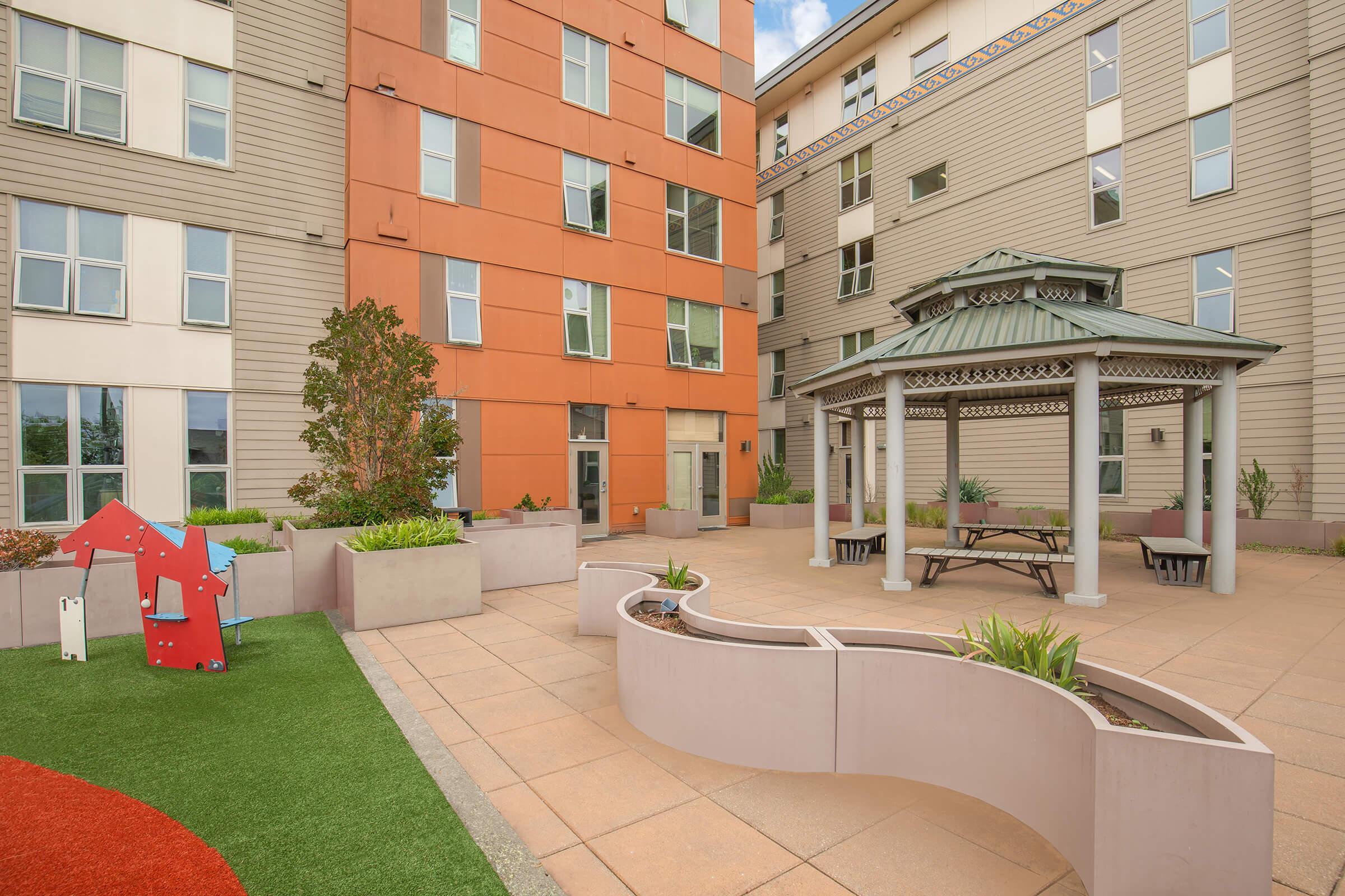 A modern courtyard featuring a landscaped area with grass and colorful play structures. In the center, a gazebo provides shade over benches and tables. Surrounding the area are contemporary buildings with orange and neutral tones, creating a welcoming community space.