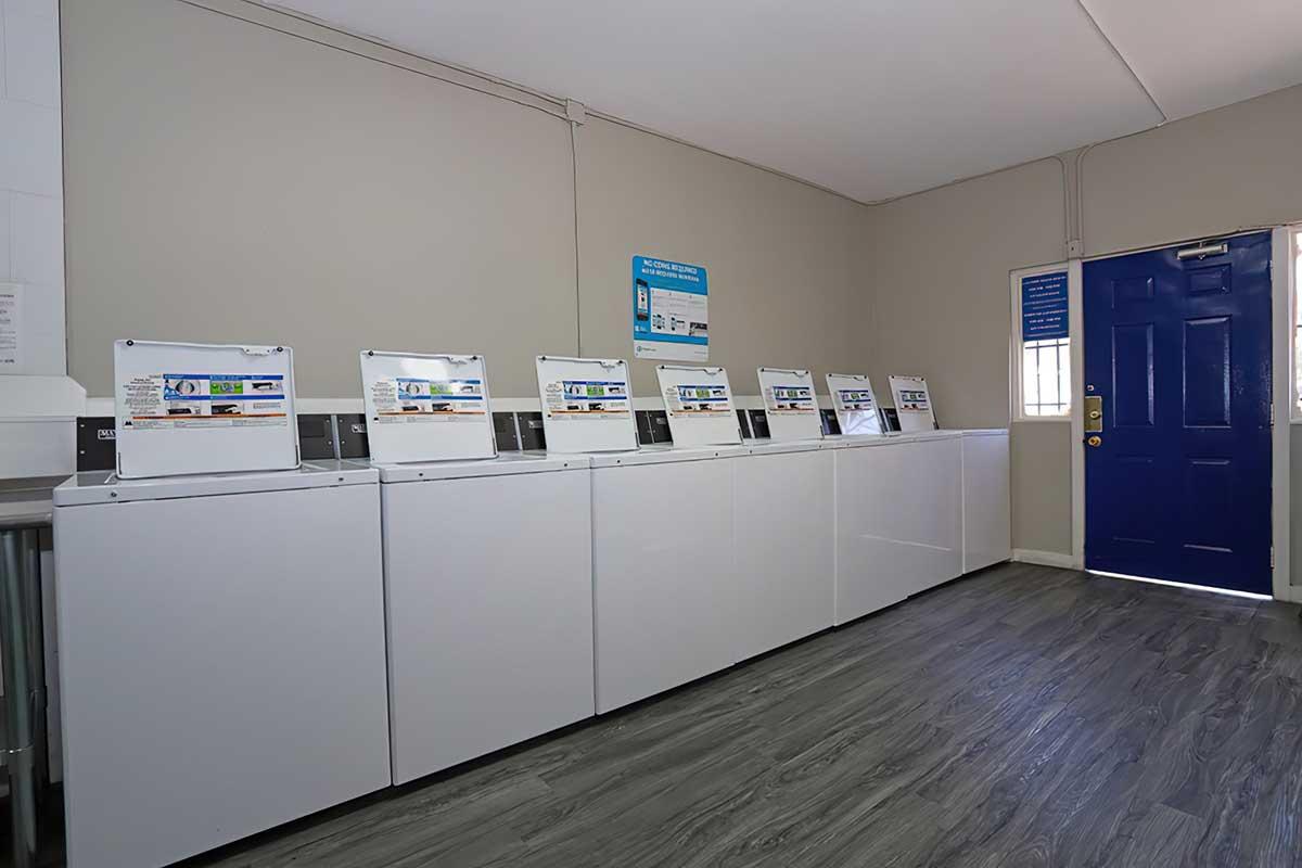 A row of white washing machines in a laundry room, positioned against a light gray wall. A blue door with a window is visible at the end of the room, along with a sign on the wall providing information about the laundry services. The floor features dark wood-like vinyl planks.