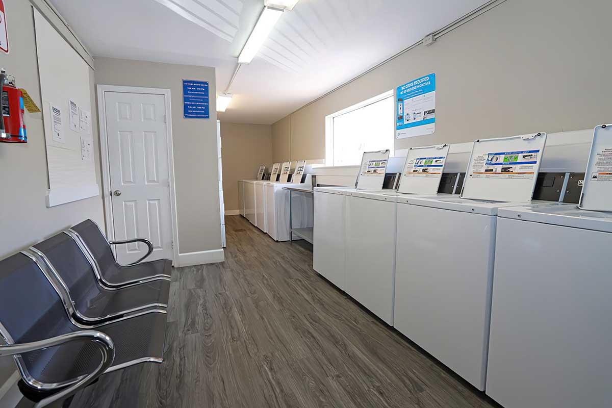 A clean and well-lit laundromat interior featuring several washing machines along one wall, a row of metal seating on the left, and informational signs on the walls. The space has a modern design with wood-like flooring and large windows providing natural light.