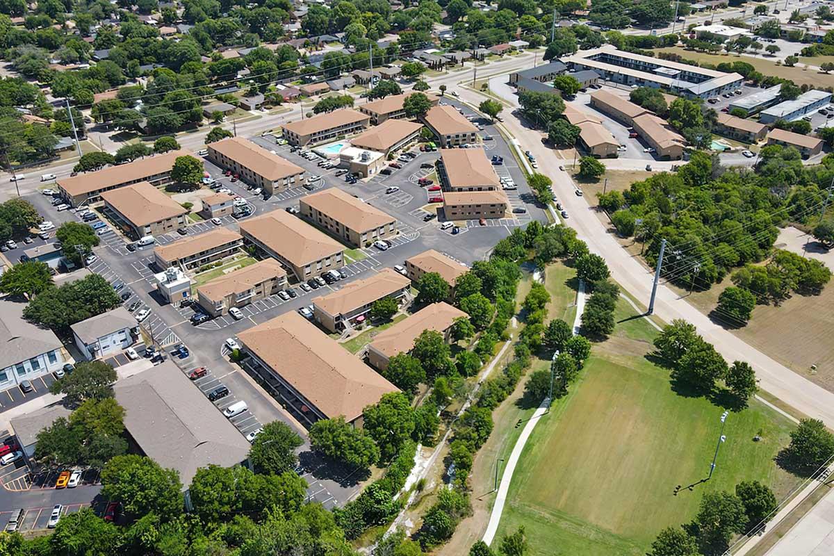 Aerial view of a residential area featuring several buildings with tan rooftops, surrounded by parking lots and trees. A green park is visible in the lower part of the image, with pathways and open grassy spaces. Nearby streets and additional buildings are also seen in the background.
