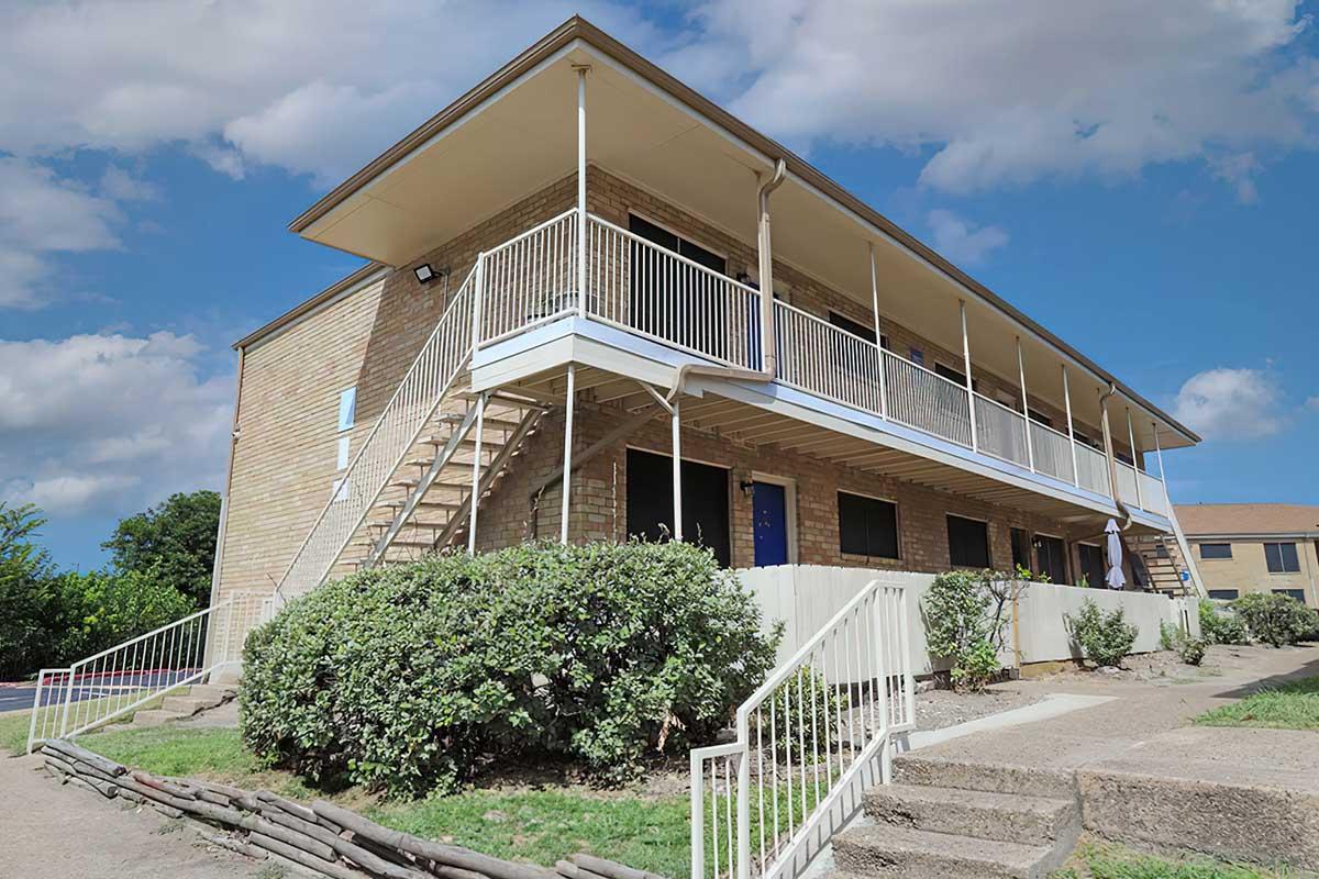 A two-story brick apartment building with multiple entrances and a staircase leading to the upper level. It features a white railing on the balcony and is surrounded by well-maintained shrubs and grass. The sky is partly cloudy, adding a bright atmosphere to the scene.