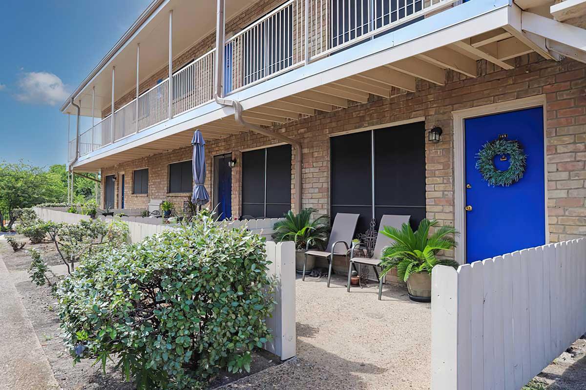 A well-maintained outdoor area of an apartment building featuring a pathway lined with plants, two chairs, and a blue door adorned with a wreath. The building has a balcony with an umbrella and large windows with screens. The clear blue sky enhances the inviting atmosphere.