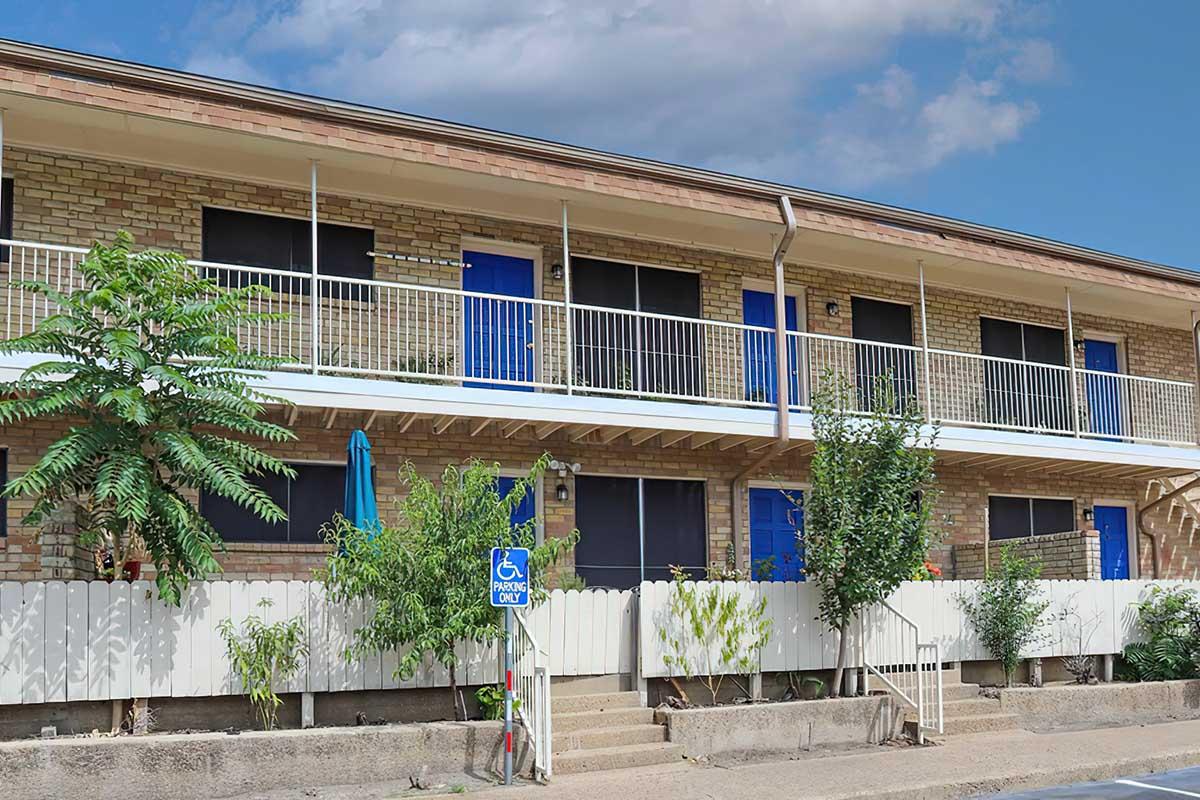 A two-story residential building with a brick facade. The upper level features blue doors and a balcony, while the ground level includes a small patio with plants. A wheelchair-accessible ramp is visible, along with a white fence and various greenery in front of the building. The sky is partly cloudy.
