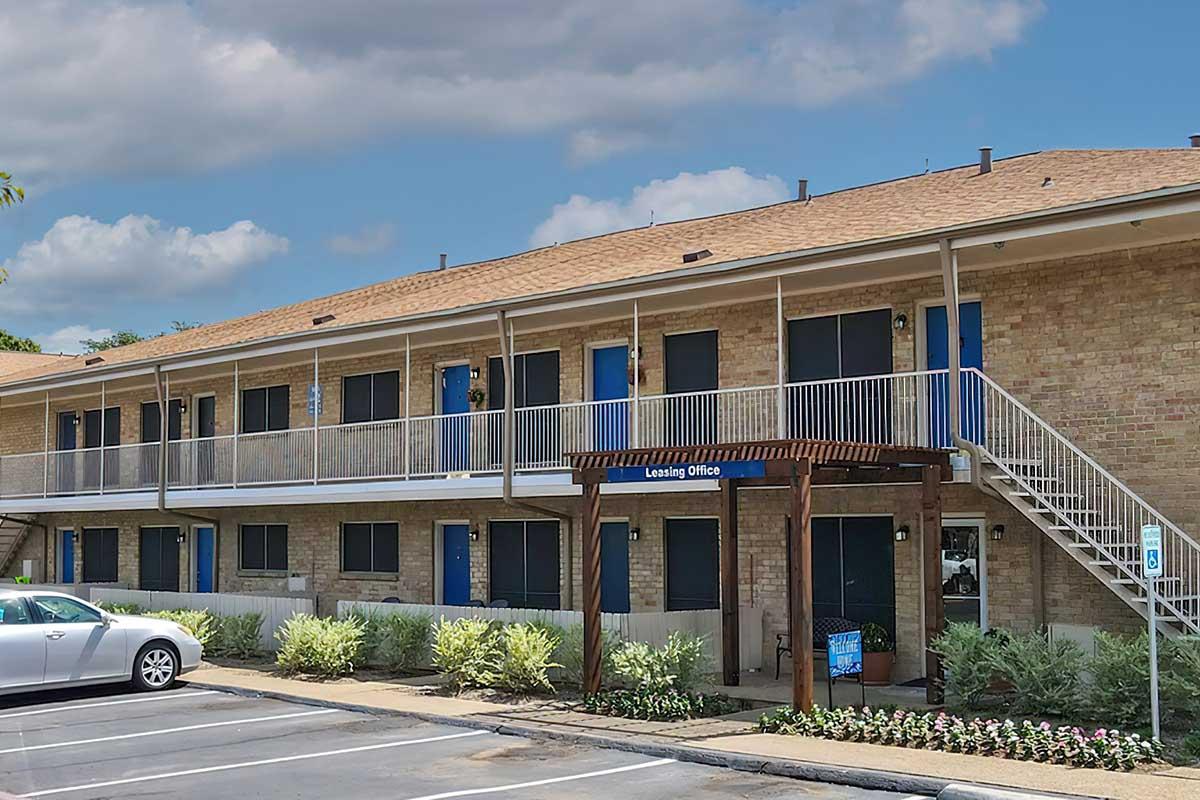 A two-story apartment building with a brick exterior, featuring a leasing office entryway. The building has blue doors on each unit and a staircase leading to the upper level. Surrounding the entrance are small shrubs and parking spaces in front of the building. The sky is partly cloudy.
