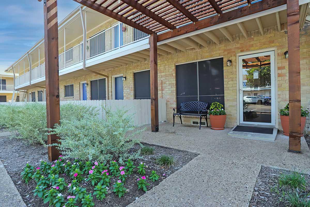 A view of a landscaped outdoor area in front of a multi-story building. There is a bench under a wooden pergola, surrounded by flower beds with pink flowers. The entrance to the building is visible, featuring a door with a small step. Soft greenery enhances the inviting atmosphere.