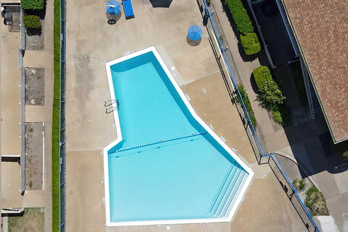 Aerial view of a uniquely shaped swimming pool surrounded by a concrete deck. The pool features a light blue color and has a shallow section with pool steps. There are blue umbrellas and lounge chairs nearby, along with green landscaping and a portion of a building visible in the background.