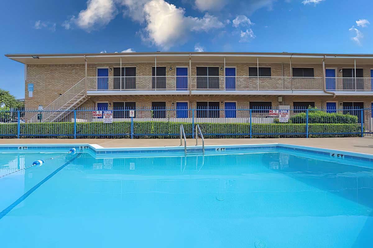 A clear blue swimming pool in the foreground with a ladder leading into it. In the background, a two-story building with several doors and blue accents. Lush green hedges line the pool area, and the sky is partly cloudy, creating a bright and inviting atmosphere.