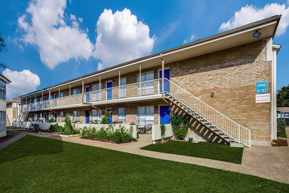 Exterior view of a two-story brick building with multiple apartments. The building features white railings on the balconies, blue doors, and a staircase on the right side. Lush green lawn and landscaping are present in the foreground, with a clear blue sky and fluffy clouds above.