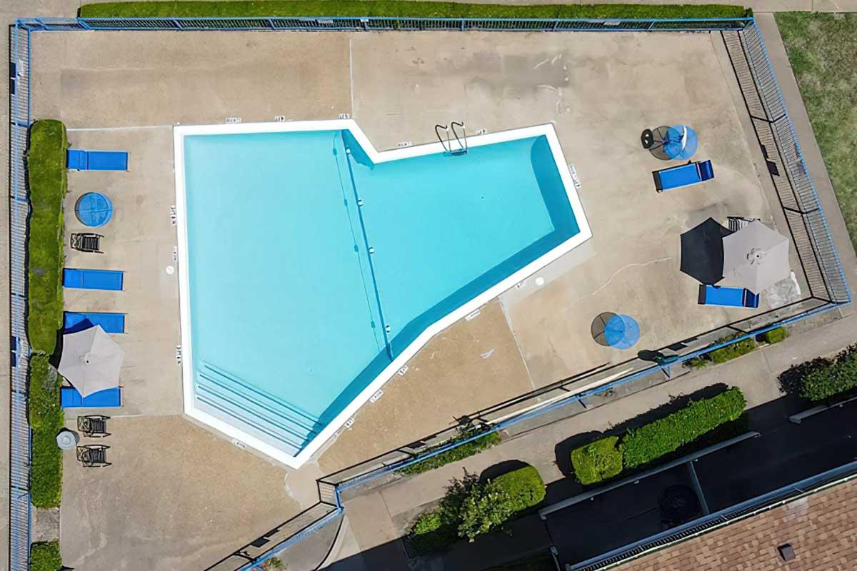 Aerial view of a uniquely shaped swimming pool surrounded by lounge chairs and tables. The pool is bright blue, with a smooth surface and marked lanes. The surrounding area includes green grass and umbrellas for shade, set in a sunny environment.