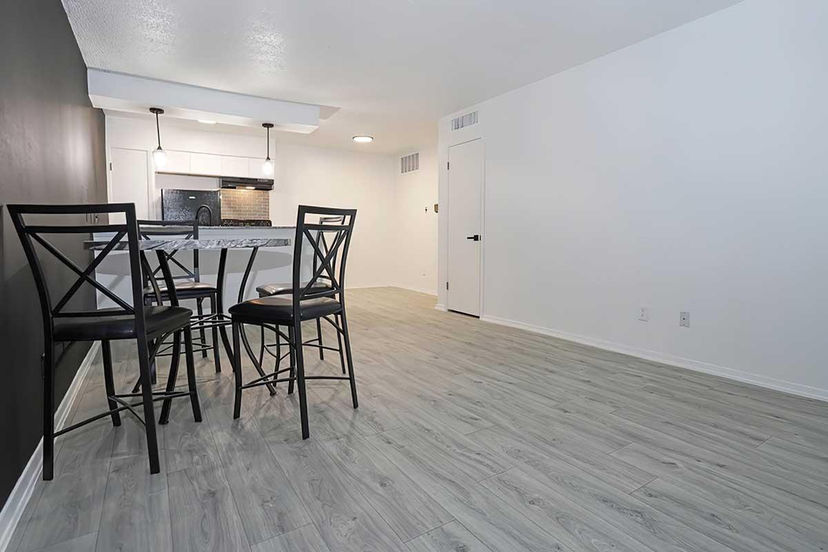 A modern, minimalistic kitchen and dining area featuring a small table with four black bar stools. The kitchen has dark countertops and white cabinets, while the walls are painted in neutral colors. The floor is light wood laminate, and there is a door on the right leading to another area.
