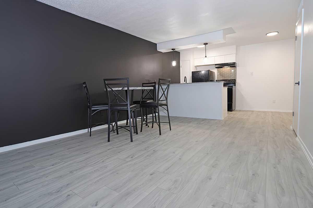 A modern, open-concept living area featuring a dining table with four black chairs, light wood flooring, and a kitchen area with sleek white cabinets and dark countertops. The wall behind the dining area is painted dark gray, adding contrast to the space.