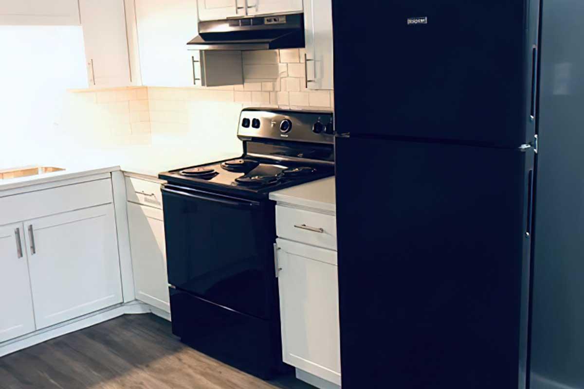A modern kitchen featuring a black stove and refrigerator. The stove sits between white cabinetry, with a sleek exhaust hood above. The countertops are minimalist, complementing the overall contemporary design of the space, which has light-colored tile backsplash and a wood-look flooring.