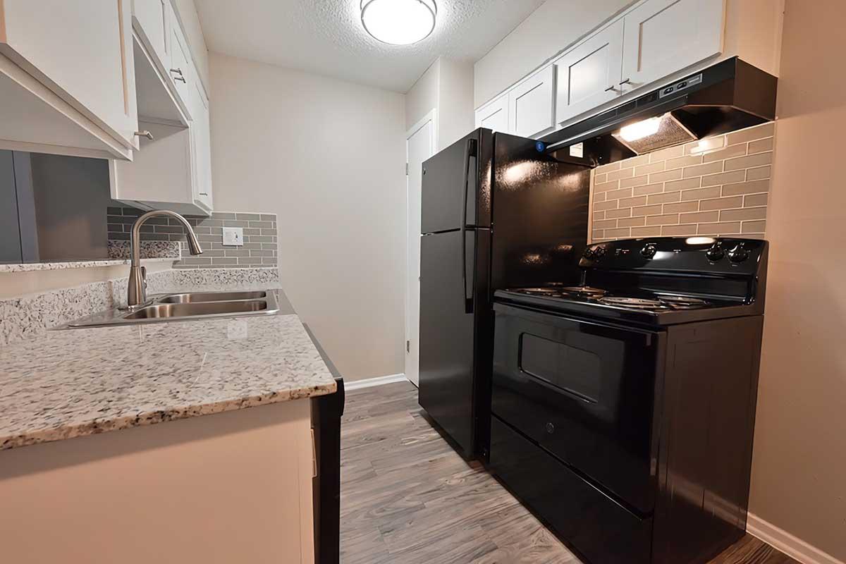 A modern kitchen featuring white cabinets, a granite countertop, and stainless steel sink. There is a black refrigerator and an oven with a matching black hood, set against a beige wall with a decorative backsplash. The flooring is light wood, contributing to a contemporary aesthetic.