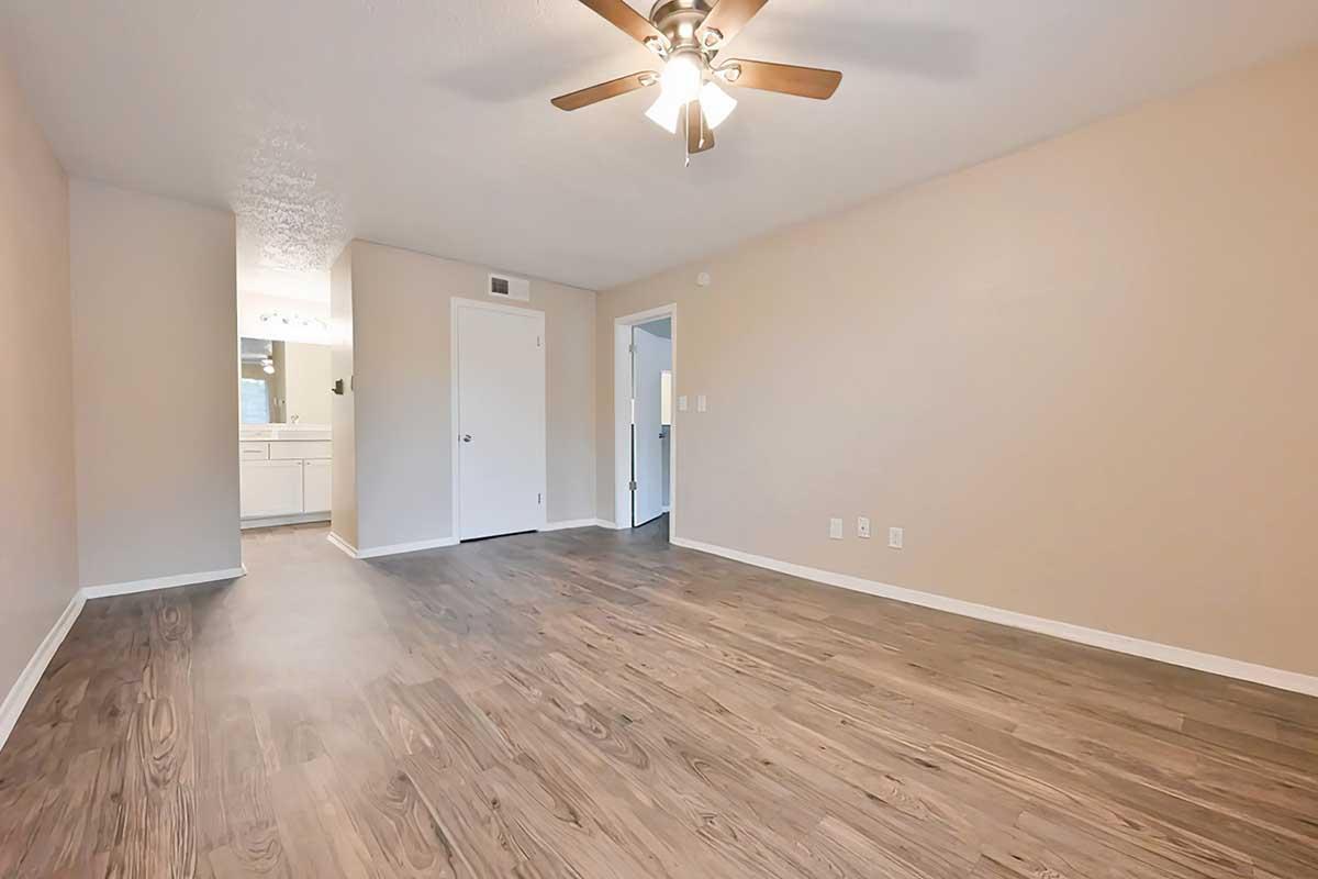A spacious, empty room with a ceiling fan and light fixture. The walls are painted a light beige, and the floor features a wood-like finish. A bathroom door and another door are visible in the background, along with a glimpse of a vanity area. The overall atmosphere is bright and open.