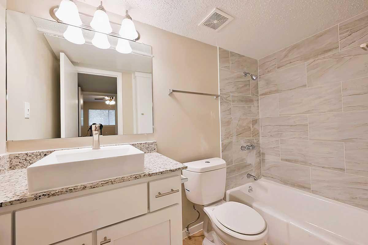 A modern bathroom featuring a beige and gray tiled design, a white sink with a granite countertop, a toilet, and a bathtub. Bright overhead lights illuminate the space, and a mirror reflects the light. Soft colors create a clean and inviting atmosphere.