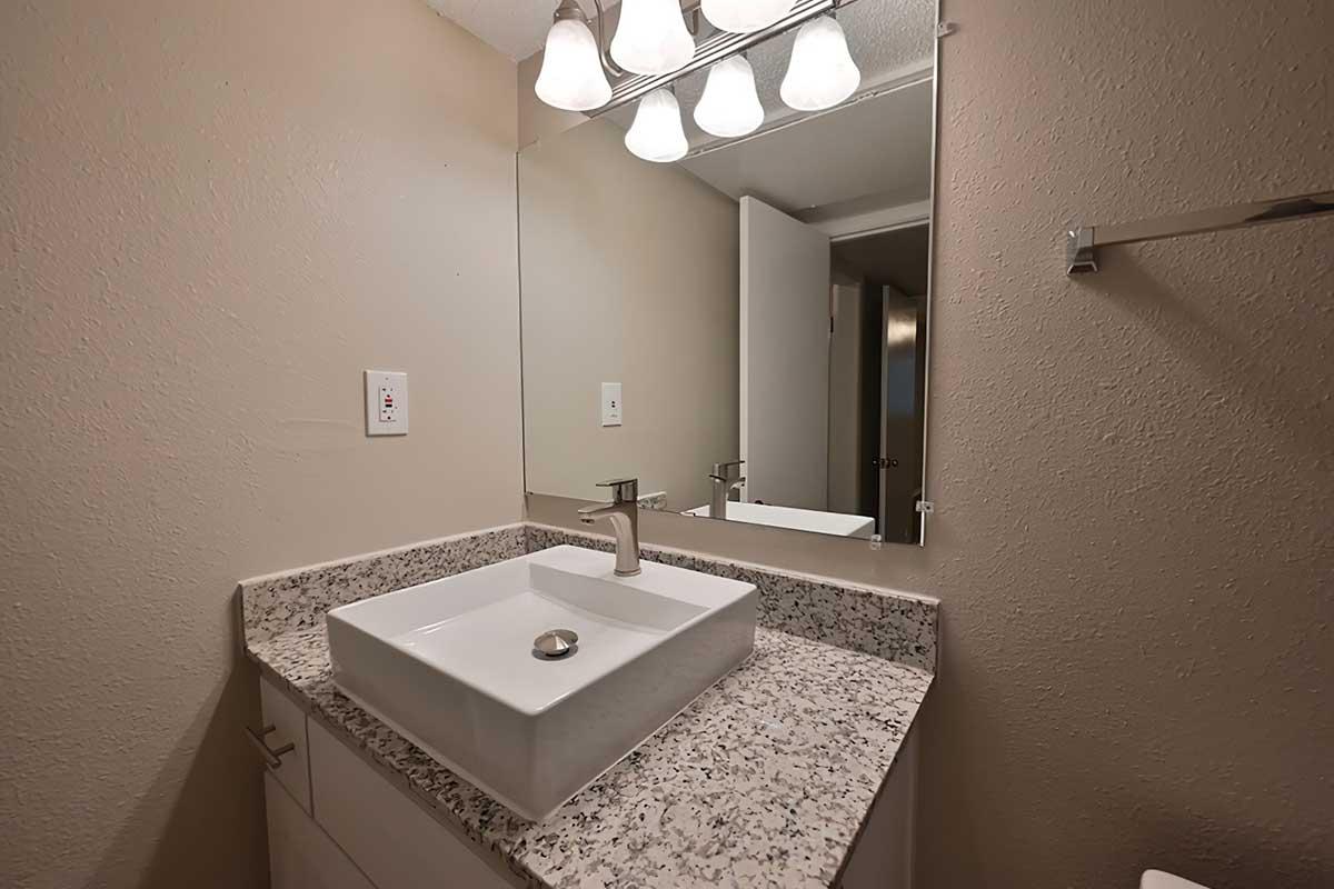 A modern bathroom featuring a sleek, white square sink set on a granite countertop. Above, a row of bright light fixtures illuminates the space, which has beige walls and a mirror reflecting the room. A partially open door is visible in the background, adding depth to the setting.