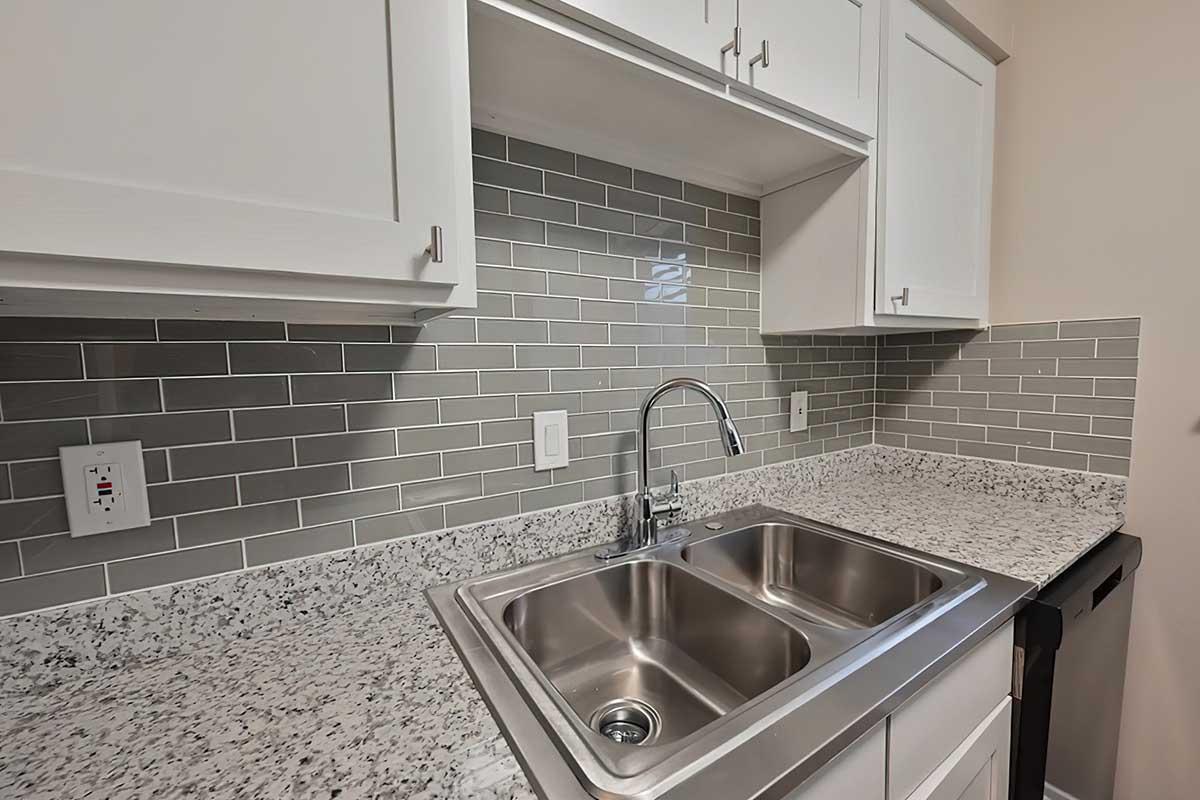 A modern kitchen featuring a double stainless steel sink with a sleek faucet, white cabinetry above, and a granite countertop. The backsplash is made of grey ceramic tiles, creating a stylish and clean look. An appliance is visible on the right.