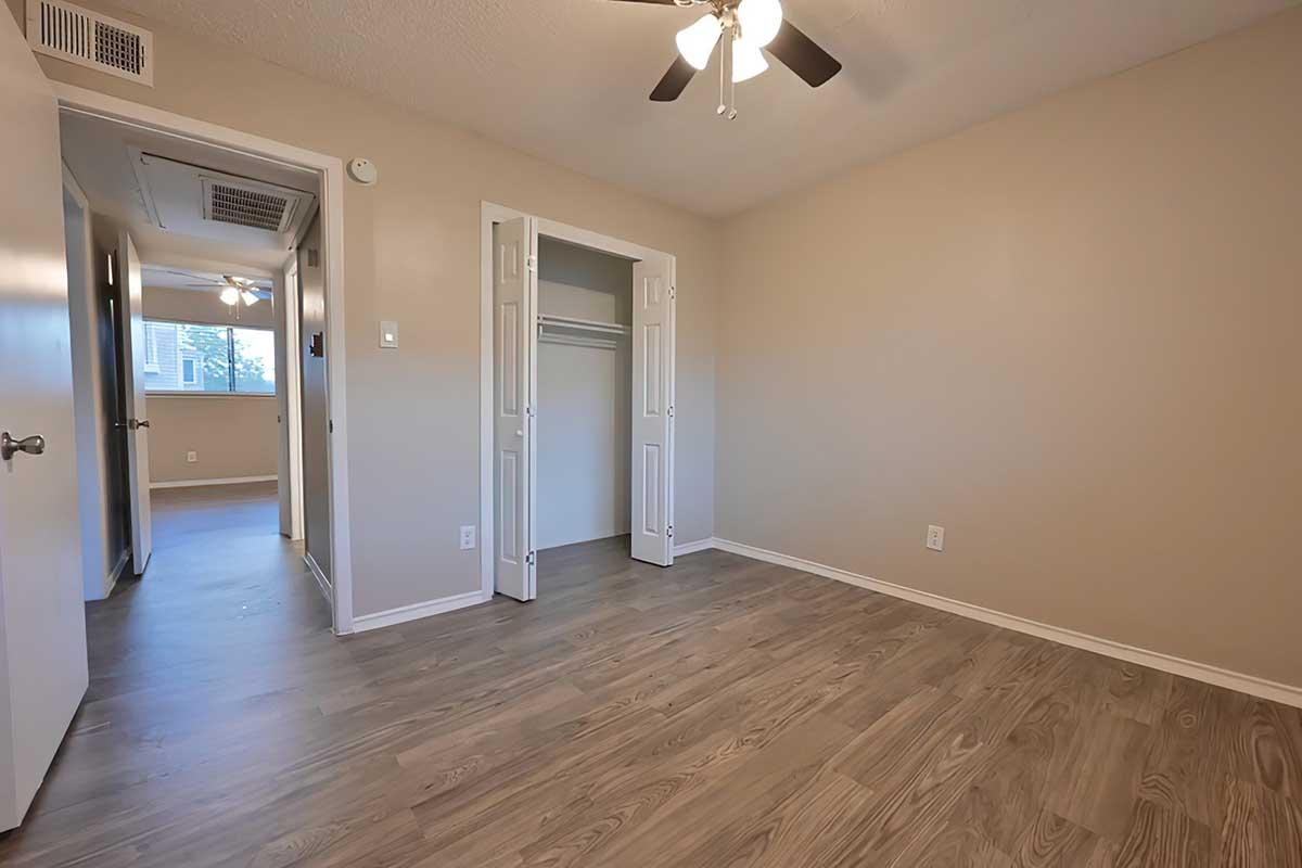A well-lit, empty room with light brown wooden flooring and beige walls. It features a ceiling fan, a closet on the right, and an open door leading to another space. The overall atmosphere is bright and inviting, ideal for a living area or bedroom.