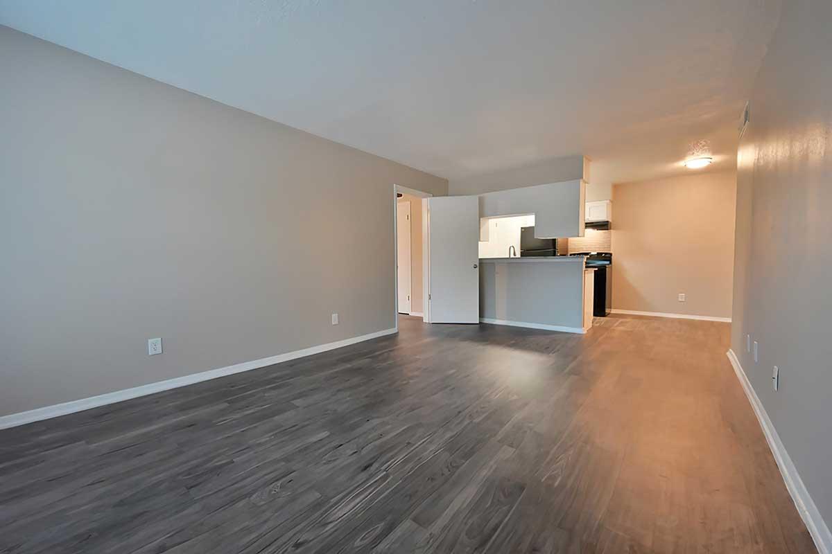 A spacious interior of a light-colored room with wooden flooring. A doorway leads to a kitchen area in the background, featuring a white wall and appliances. The space is well-lit, showcasing an open layout with neutral tones, making it suitable for various uses.