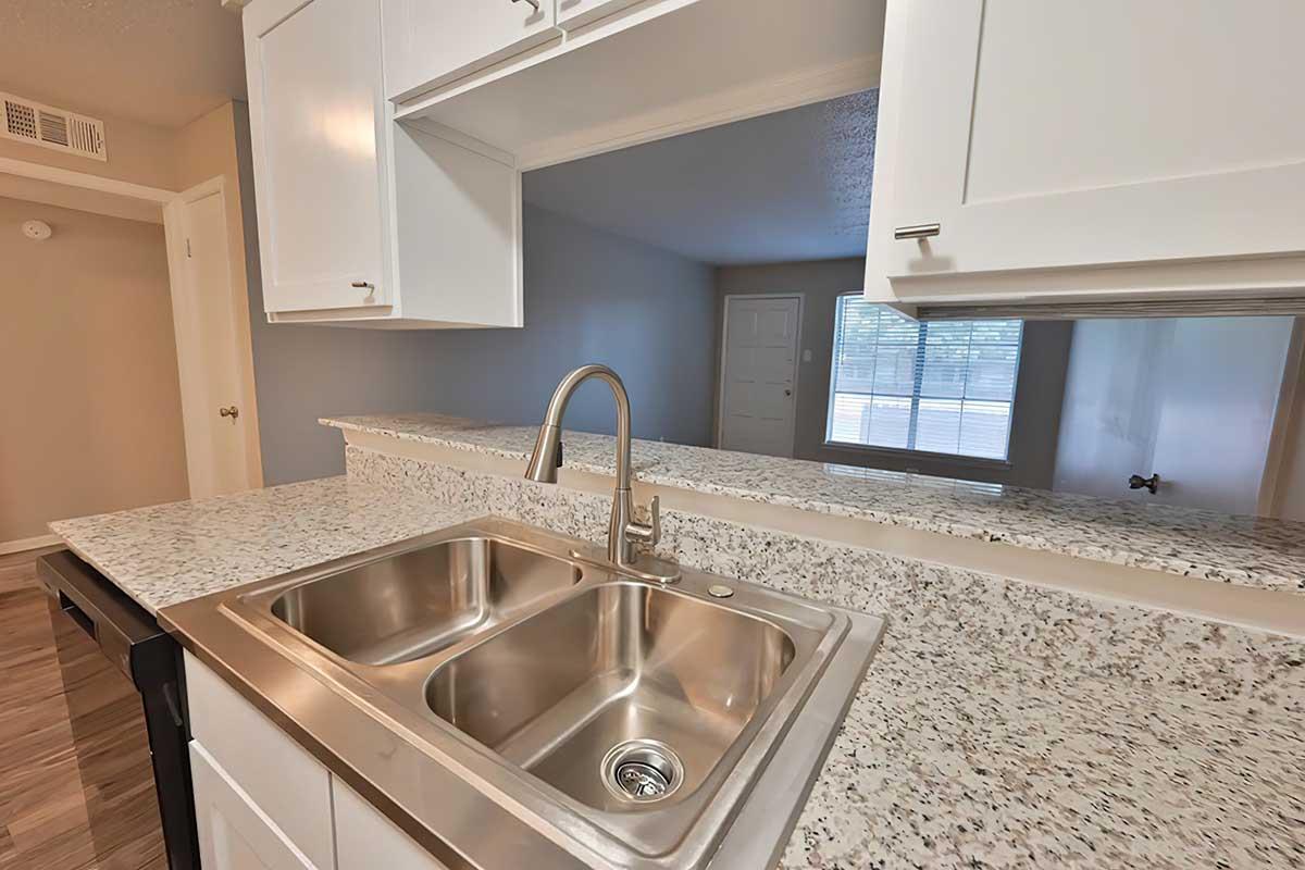 A modern kitchen featuring a double sink with a polished stainless steel finish, set against a speckled granite countertop. White cabinetry lines the walls, and a large window allows natural light to illuminate the space, with a view of another room in the background.