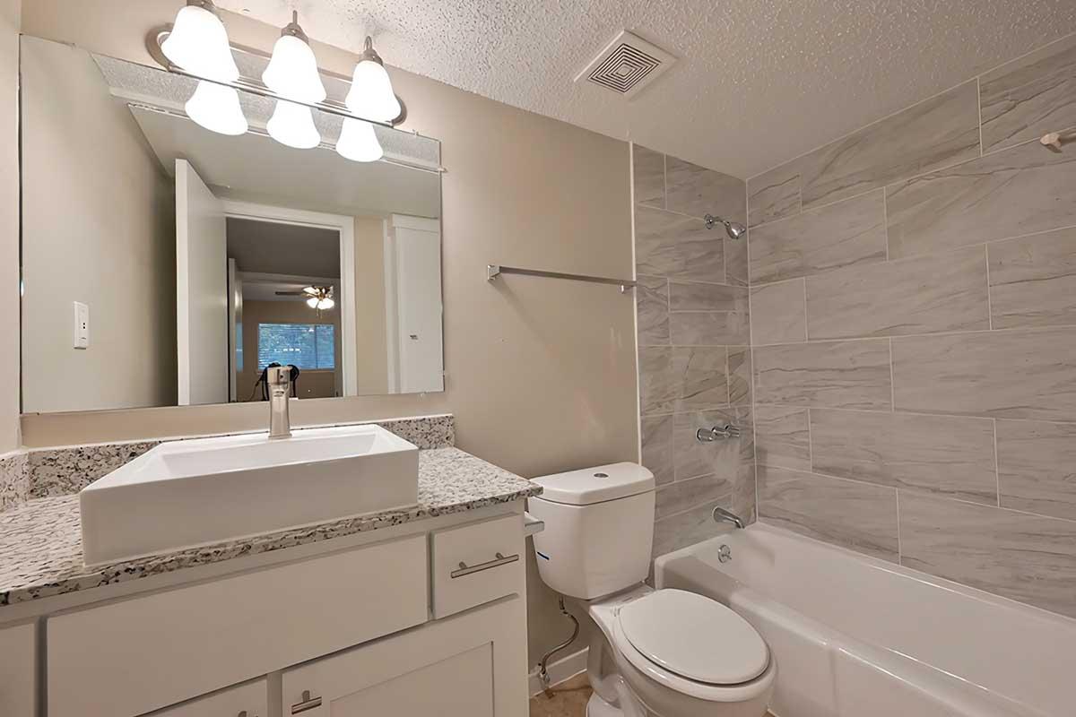 A modern bathroom featuring a white sink on a granite countertop, a wall-mounted mirror, a white toilet, and a bathtub. The walls are tiled with a light gray stone pattern, and there are bright overhead lights providing illumination. A door is visible in the background leading to another room.