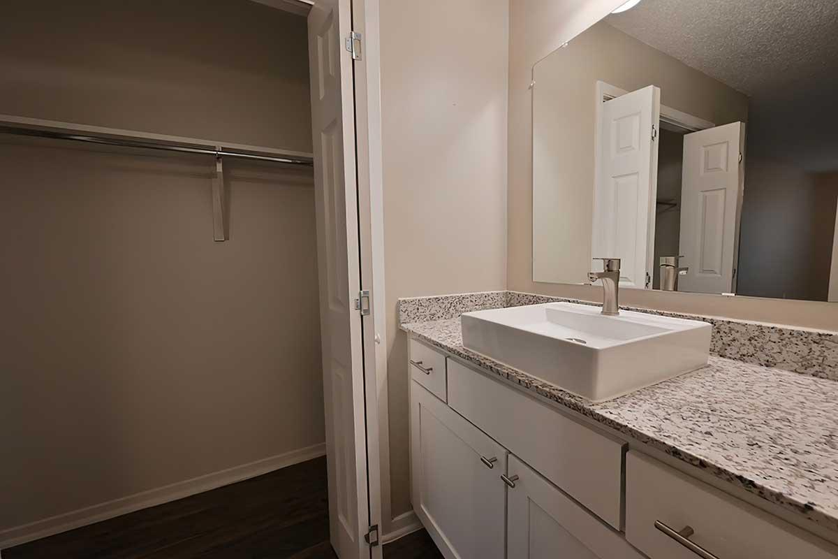 A modern bathroom featuring a white sink on a granite countertop, with a closet space visible in the background. The walls are painted in a neutral tone, and there is overhead lighting. The floor appears to be dark wood. The overall design is clean and minimalistic.