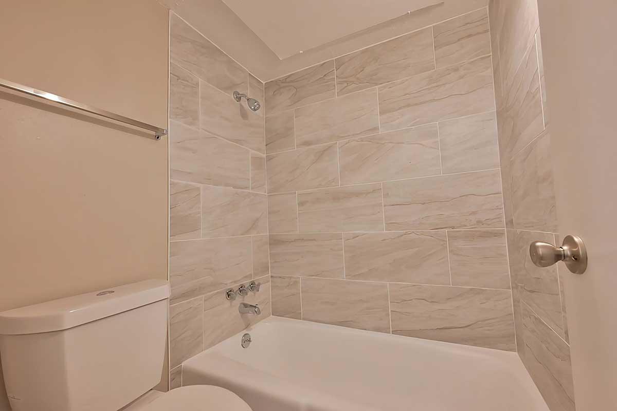 A clean bathroom featuring a bathtub with a showerhead, modern fixtures, and beige tiled walls. The toilet is positioned next to the bathtub, complementing the neutral color scheme of the space. A towel rack is visible on the wall, adding functionality to the design.