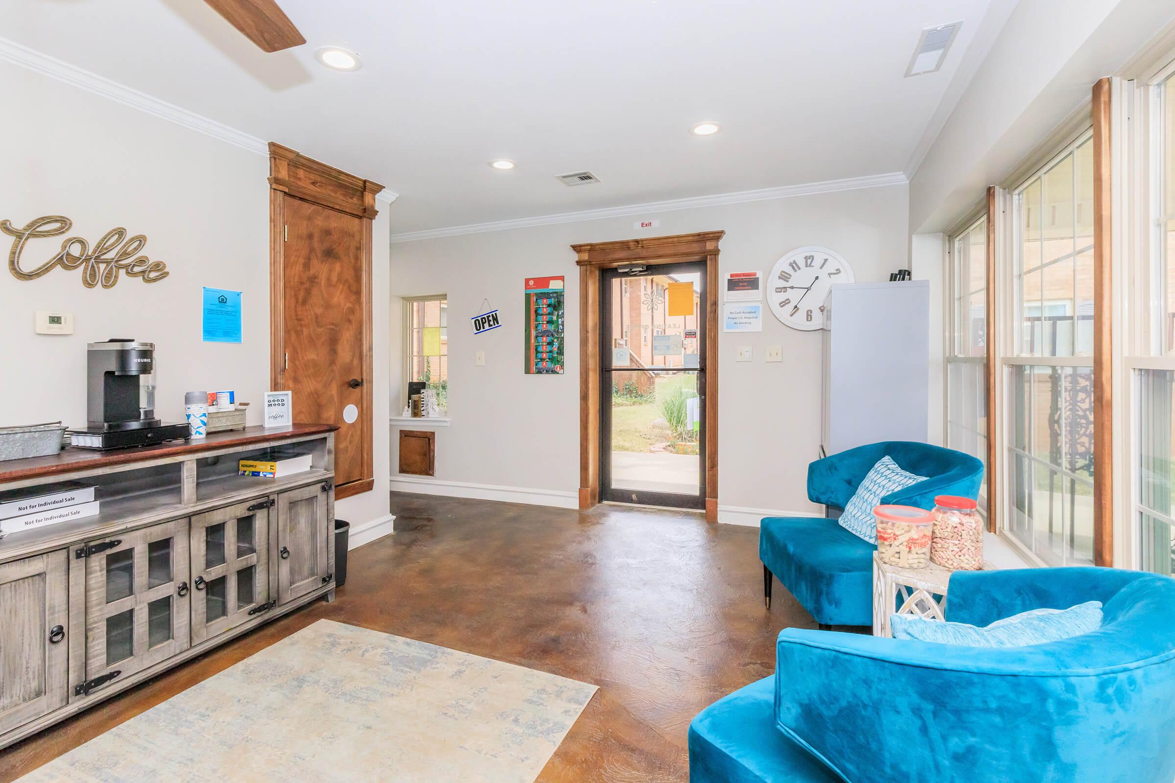 A cozy interior room featuring a coffee station with a wooden cabinet, a seating area with two blue chairs, a small table, and a decorative rug. Natural light floods in through windows, and a door leads to an outdoor space. Wall decorations include a clock and various informational flyers.