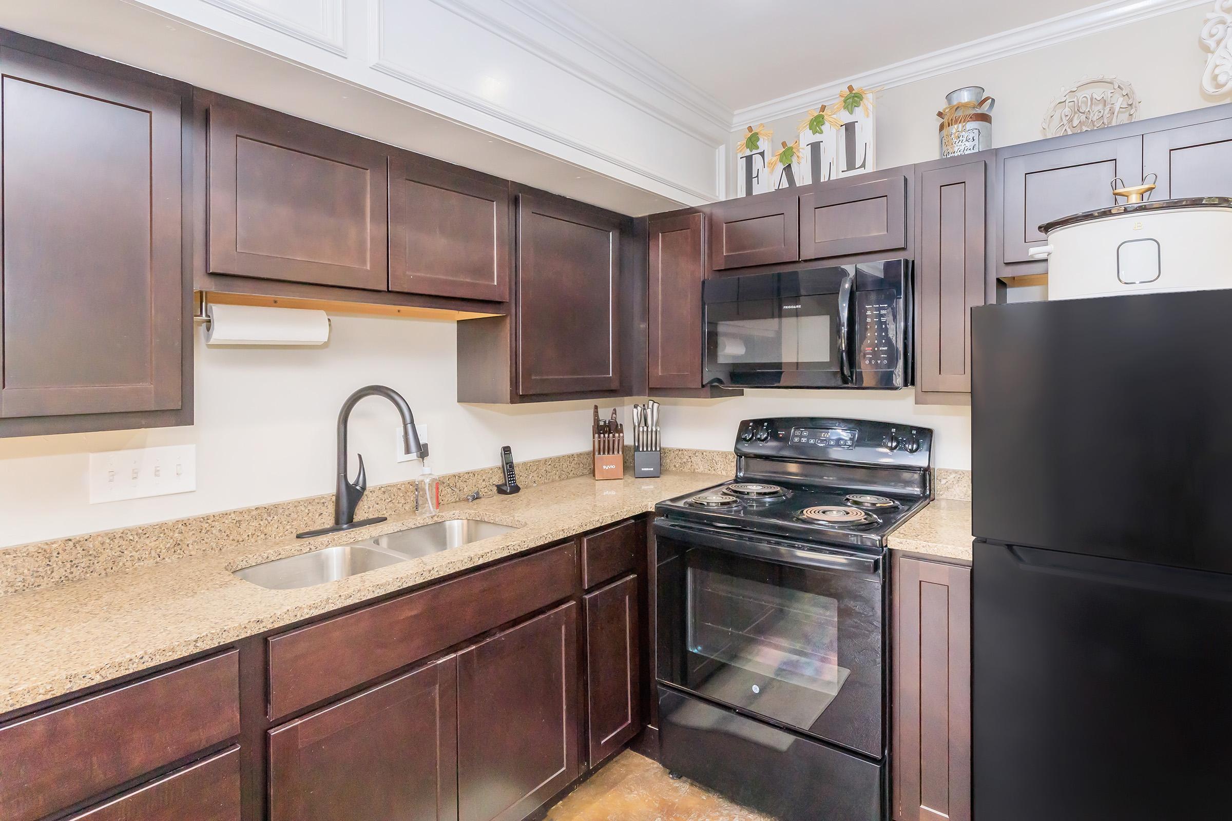 A modern kitchen featuring dark wood cabinets, a granite countertop, a stainless steel sink, and a black stove and refrigerator. The kitchen is well-organized with utensils and decorative elements on shelves, creating a clean and inviting atmosphere.