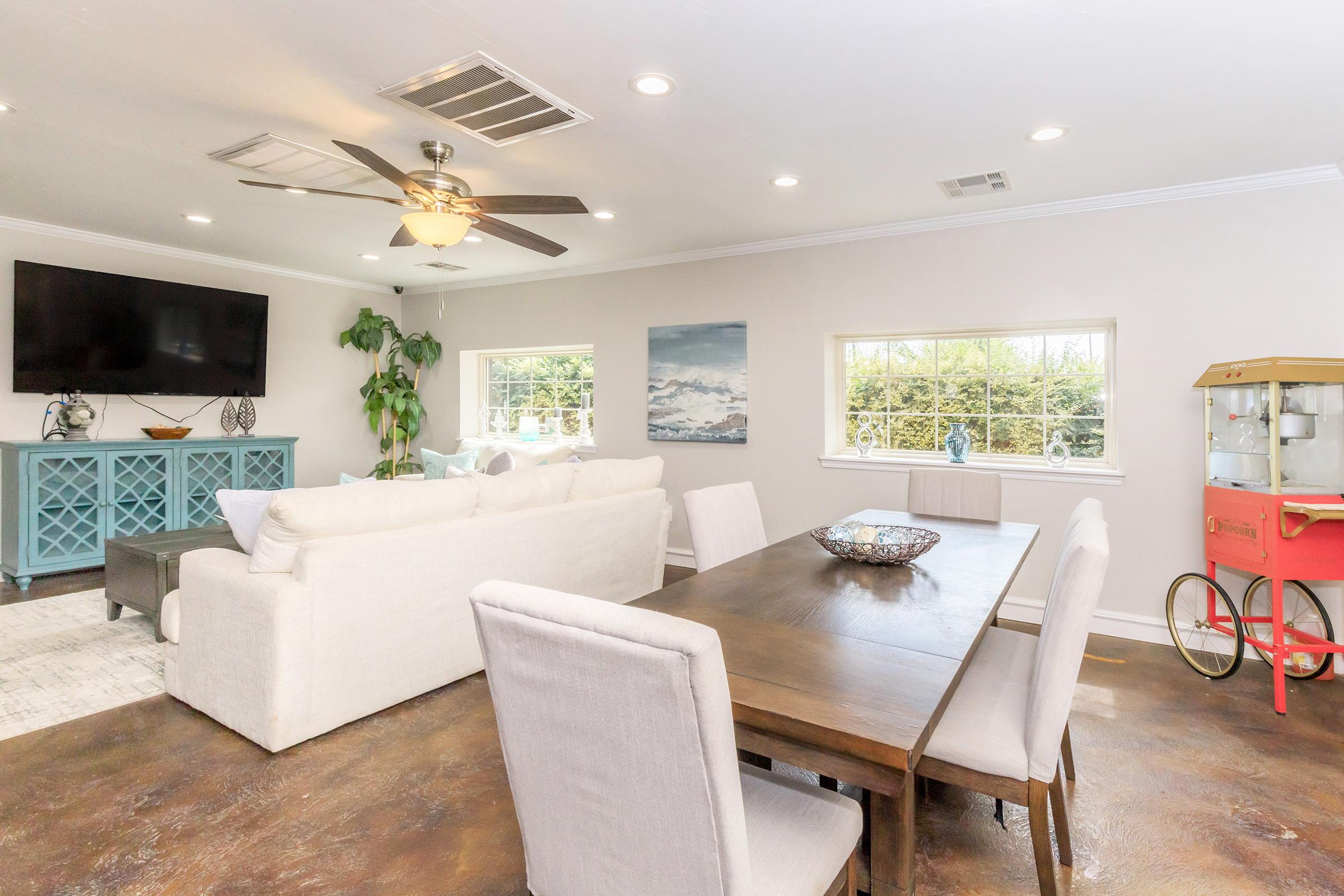 Spacious living and dining area featuring a light-colored sofa, a wooden dining table with upholstered chairs, a blue sideboard, and a vintage popcorn machine. Large windows provide natural light, and there’s a mounted TV on the wall, along with decorative plants and artwork.