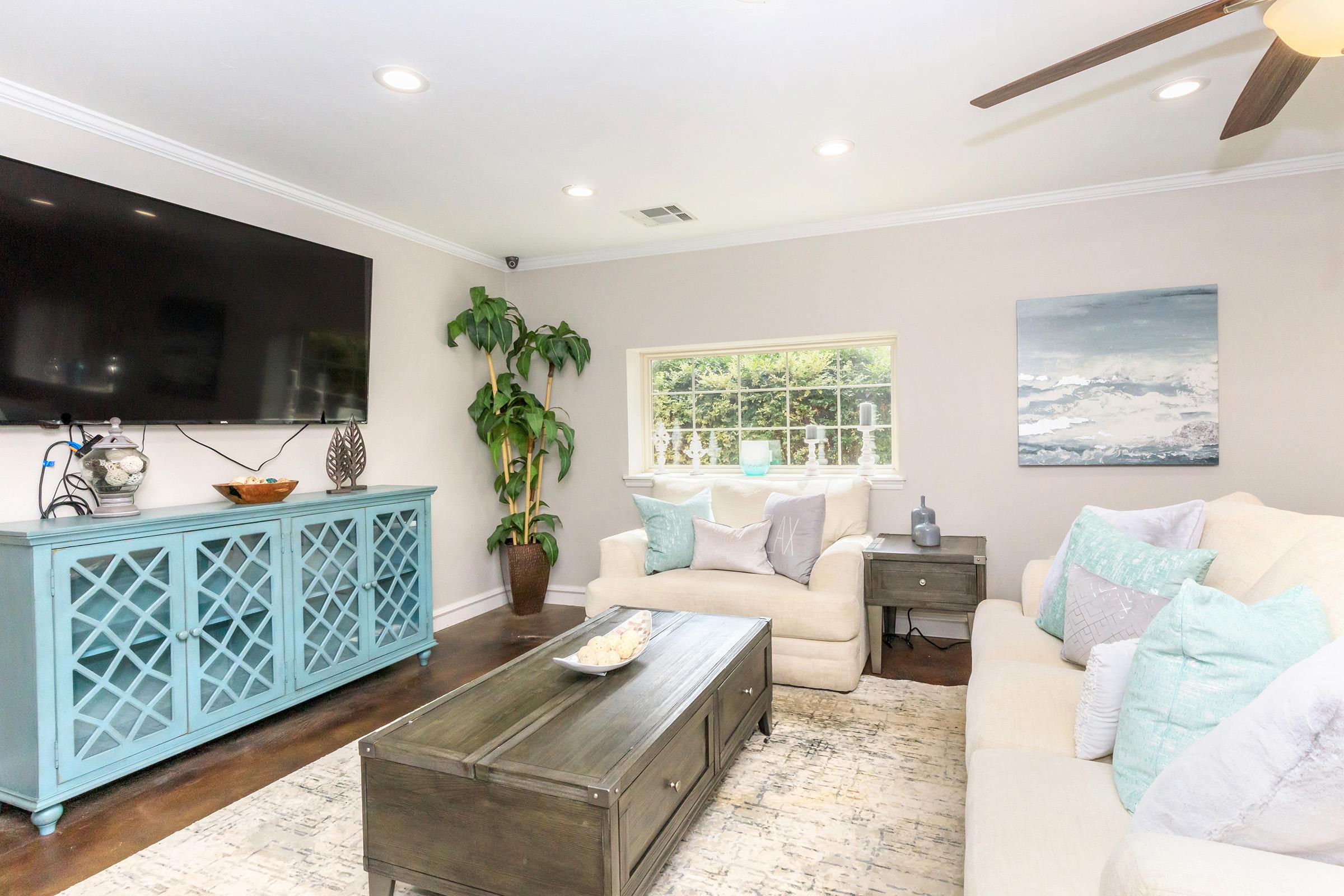 A bright and inviting living room featuring a large flat-screen TV on the wall, a light-colored sofa with decorative pillows, a blue wooden console table, and a coffee table. Natural light streams in through a window adorned with greenery, creating a cozy atmosphere. The walls are painted in a soft beige tone.