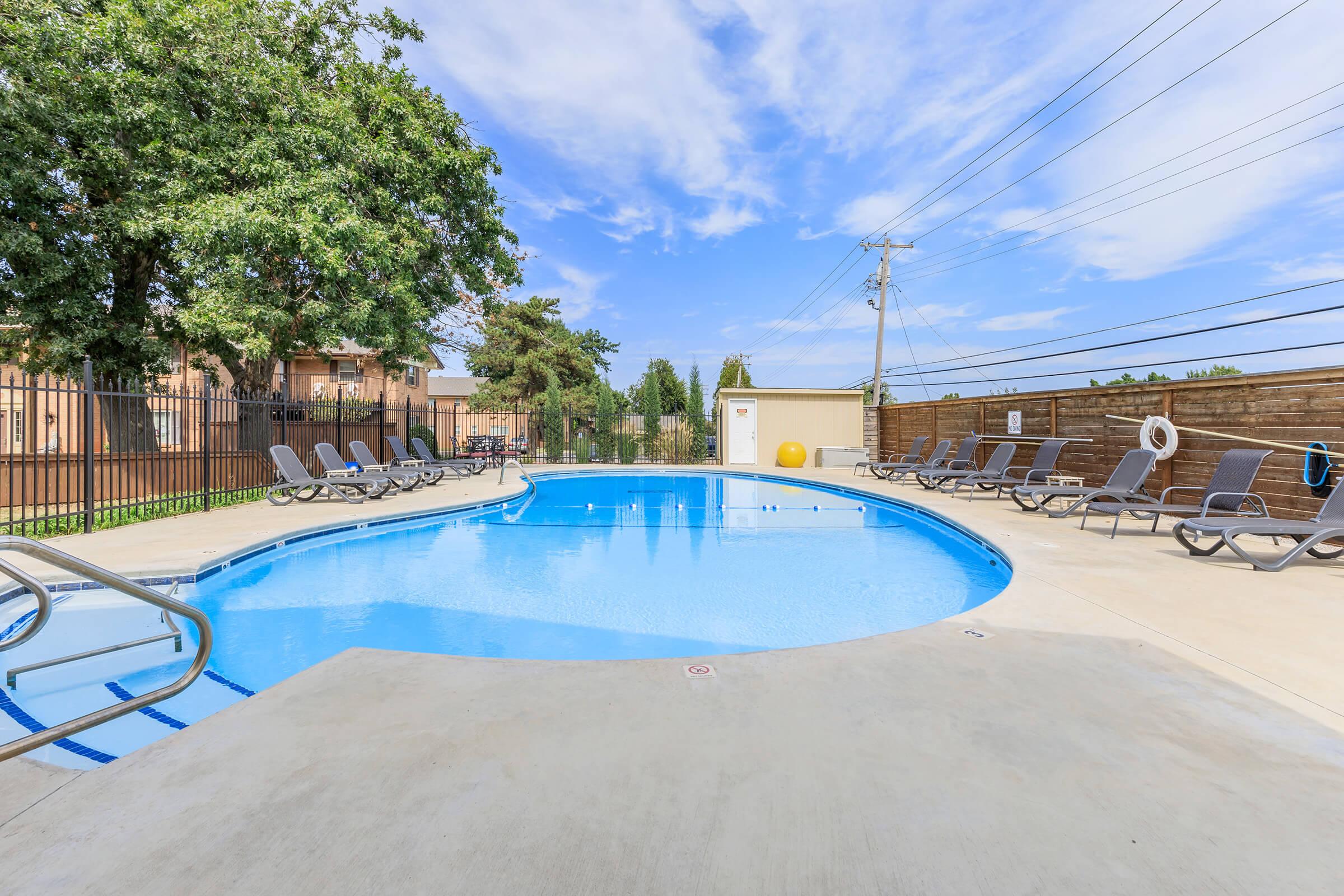 A clear blue swimming pool surrounded by a concrete deck, lounge chairs, and trees. The area is fenced with a wooden barrier, and power lines are visible in the background. The sky is bright with some fluffy clouds, creating a sunny atmosphere.