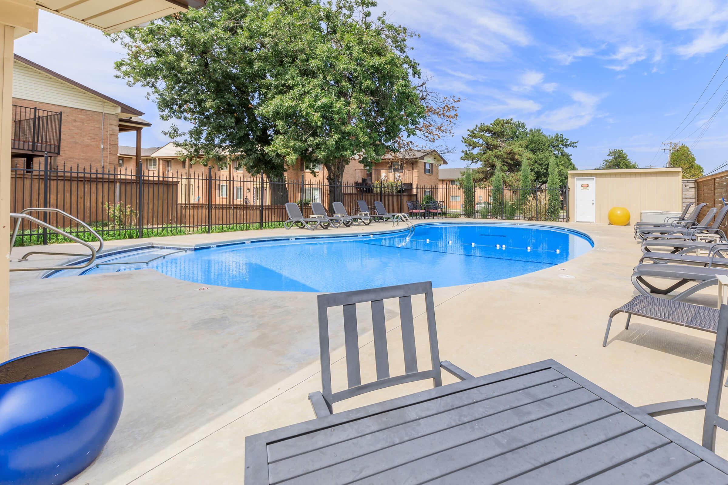 A residential pool area featuring a blue swimming pool surrounded by lounge chairs. In the background, there are apartment buildings and a large tree providing shade. A table and chairs are nearby, creating a relaxing outdoor space.
