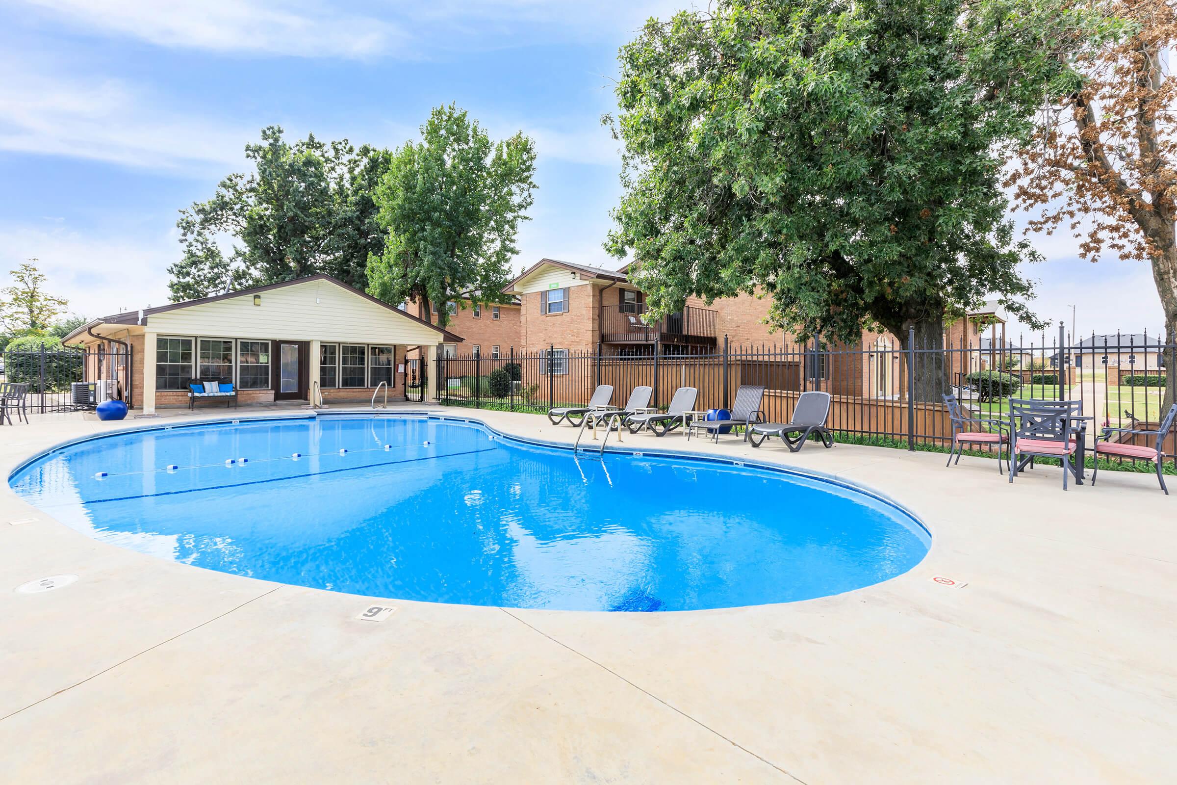 A clear blue swimming pool surrounded by lounge chairs and a fenced area, with a brick building and trees in the background. The scene is sunny, creating a vibrant and inviting atmosphere.