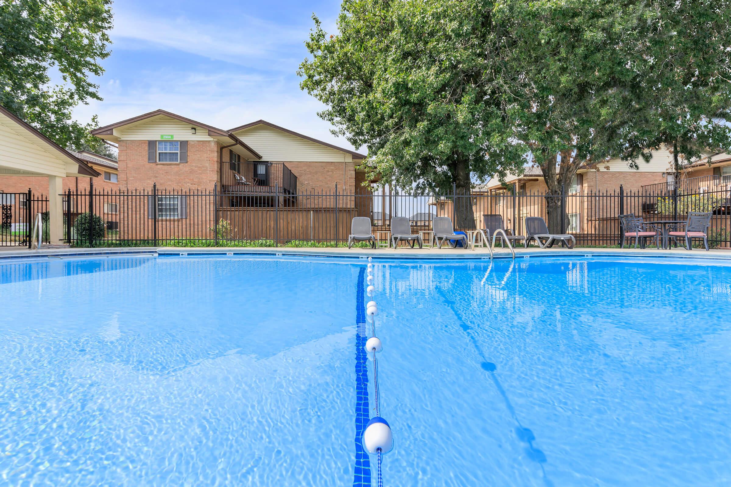 A clean, blue swimming pool surrounded by water, with lounge chairs on the deck. In the background, several residential buildings are visible, along with a black fence and trees providing shade. The sky is clear with a few clouds.