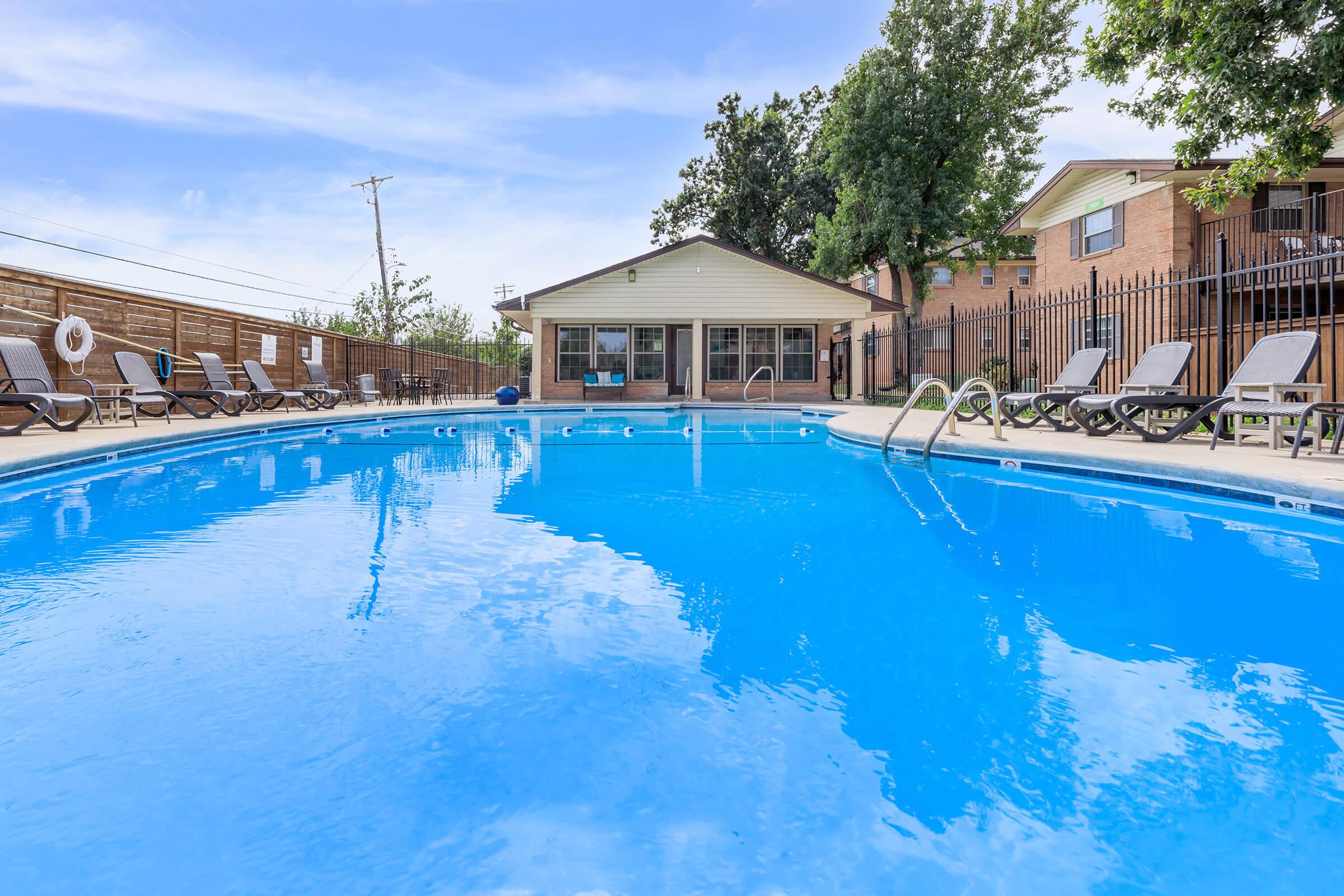 A clear blue swimming pool with lounge chairs nearby. In the background is a single-story building with large windows, surrounded by a fence and trees. The scene is bright and inviting, suggesting a relaxing outdoor space.