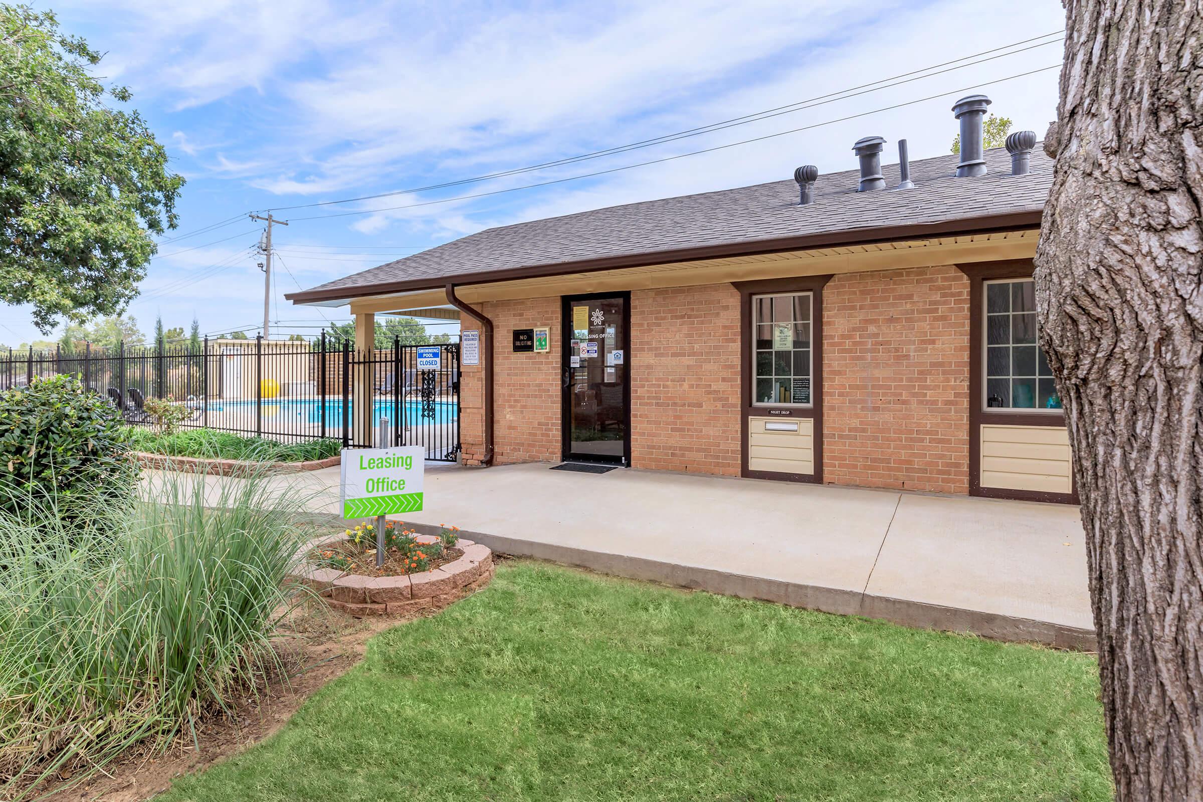 A brick leasing office building with a sign that reads "Leasing Office" in front. The entrance features large windows, and a well-maintained lawn with ornamental grasses. In the background, there's a swimming pool surrounded by a fence, under a clear blue sky with scattered clouds.