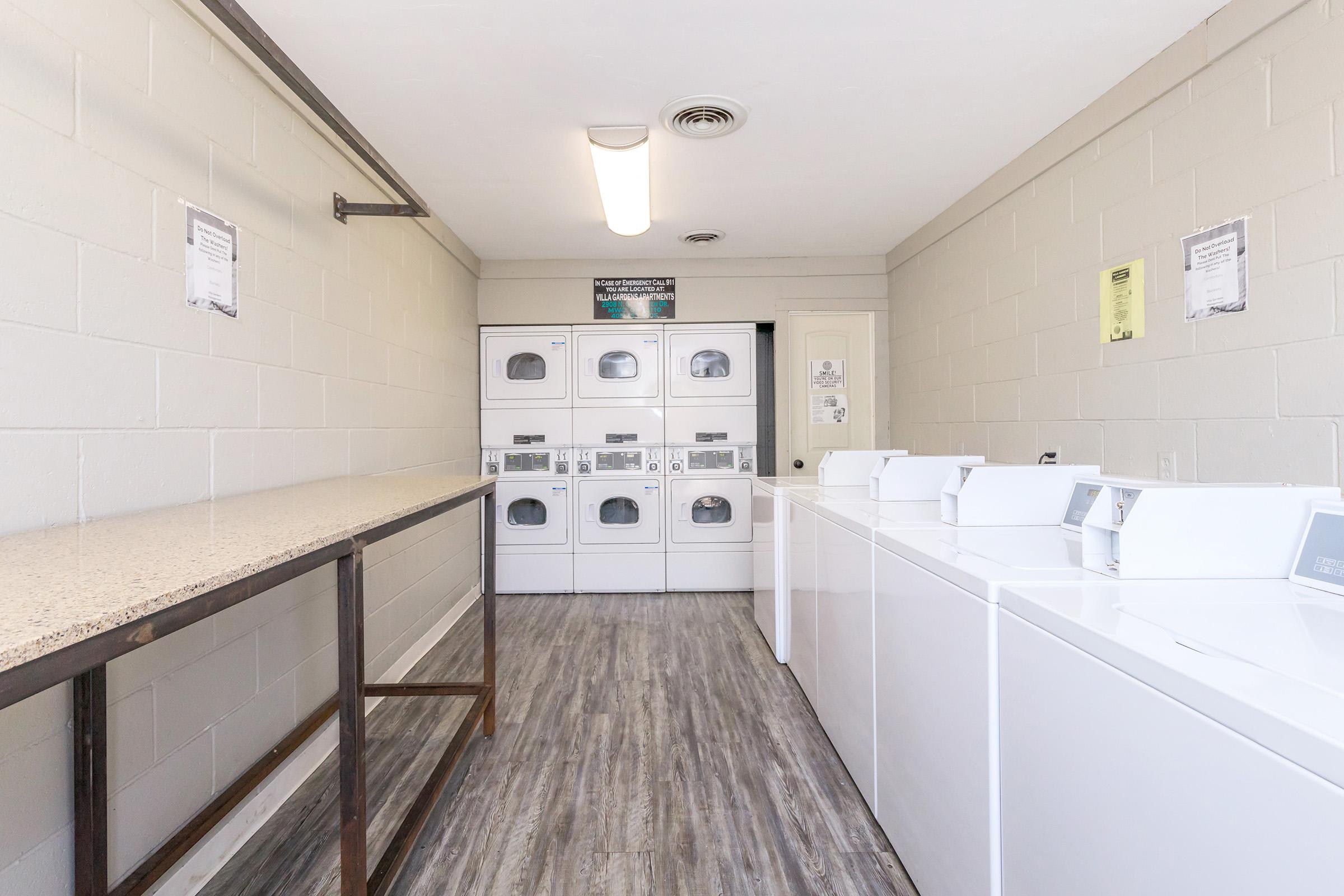 A clean and modern laundry room featuring several stacked white washing machines and dryers against a light gray wall. A long table made of wood runs alongside the machines. The space is well-lit with overhead lighting and has a minimalistic design, creating an organized atmosphere for doing laundry.